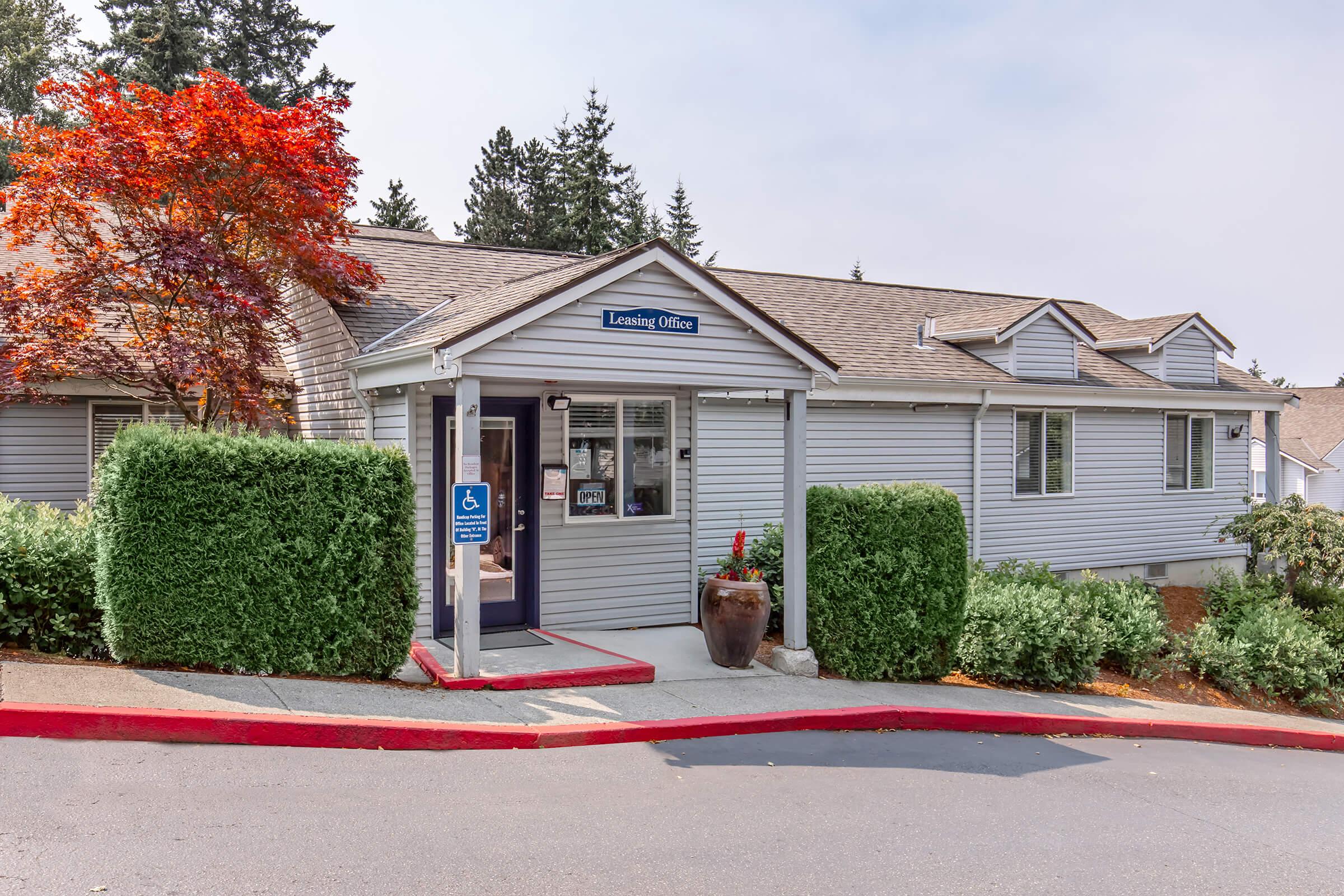 A single-story leasing office with a sign reading "Leasing Office" is situated amidst greenery. The office features large windows, a wheelchair-accessible entrance, and a landscaped area with a decorative plant. In the background, trees are visible, indicating a residential setting.