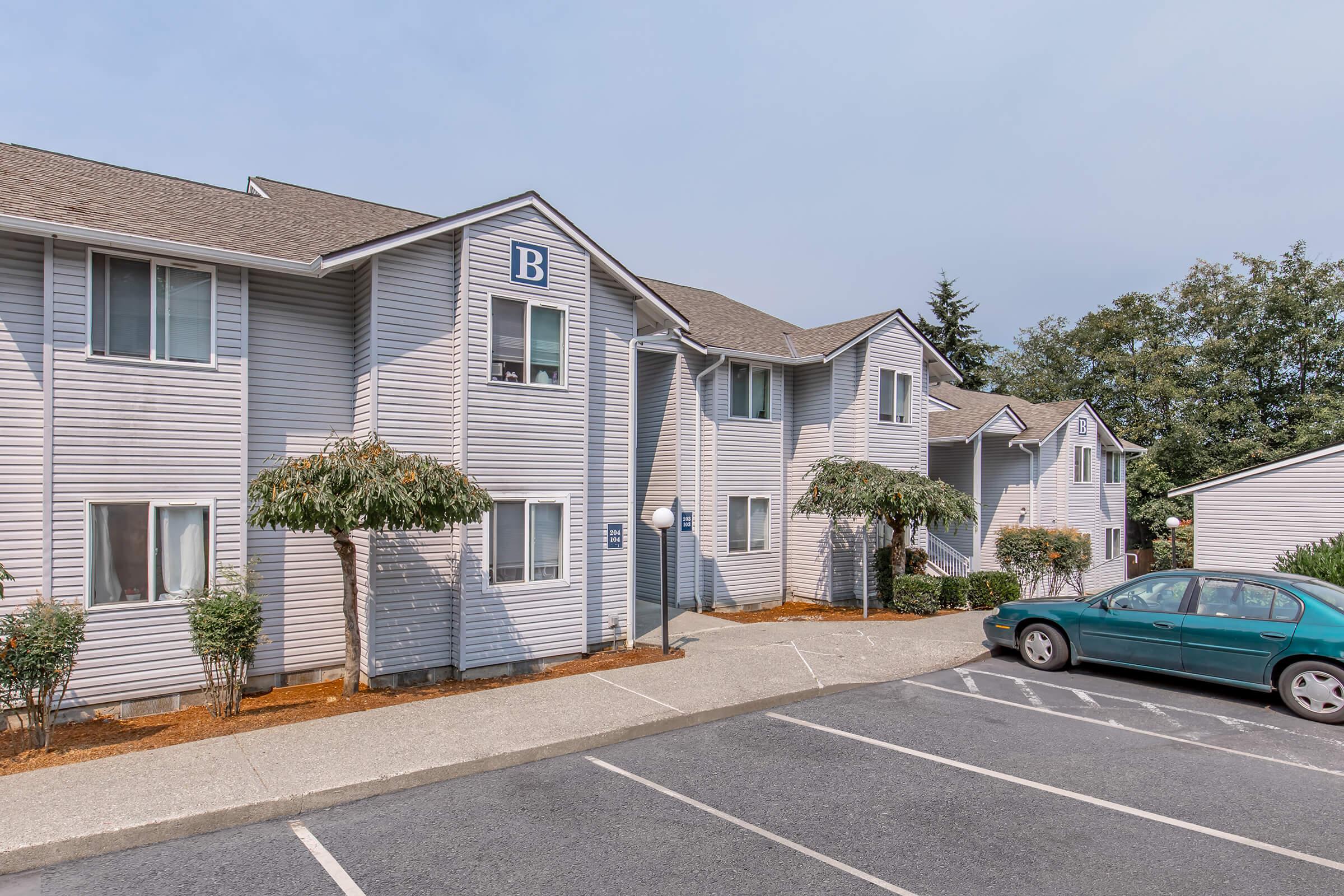 A two-story residential building with a light gray exterior, labeled "B" on the second floor. The building features multiple windows, small landscaped areas with shrubs, and a paved parking lot with a parked green car. The sky is overcast, and there are trees in the background.