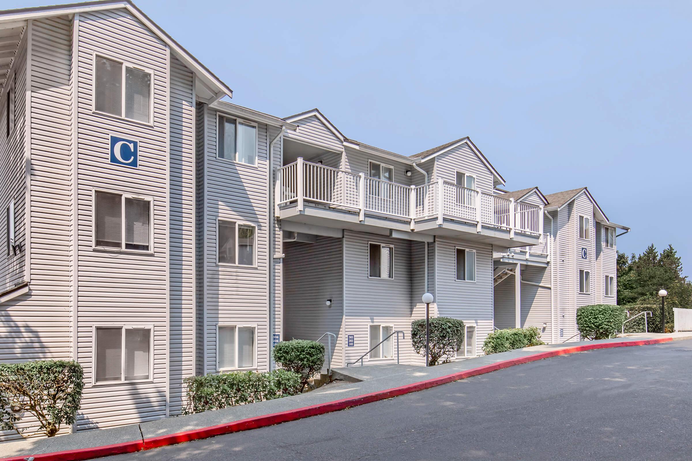 Exterior view of a multi-story residential building with a modern design. The building features multiple balconies, large windows, and a neutral color palette. A sloped driveway leads to the entrance, surrounded by neatly trimmed bushes and a red curbed area. The sky is clear, indicating a bright day.
