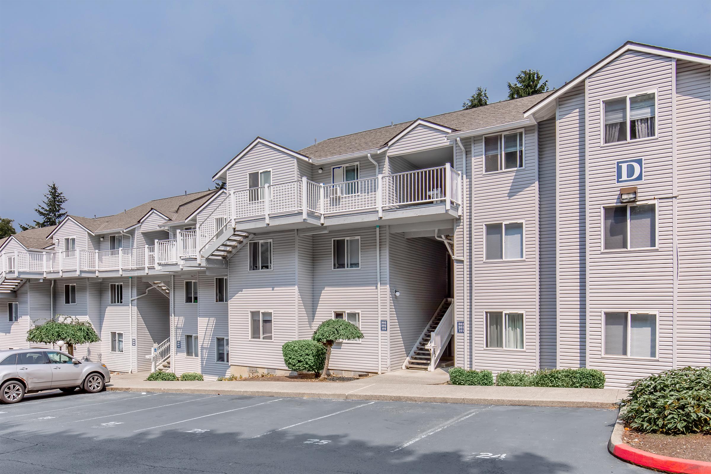 A multi-story apartment building featuring several balconies and staircases. The exterior is light gray with white trim, and a parked car is visible in the foreground. Lush green shrubs are neatly arranged near the building entrance.