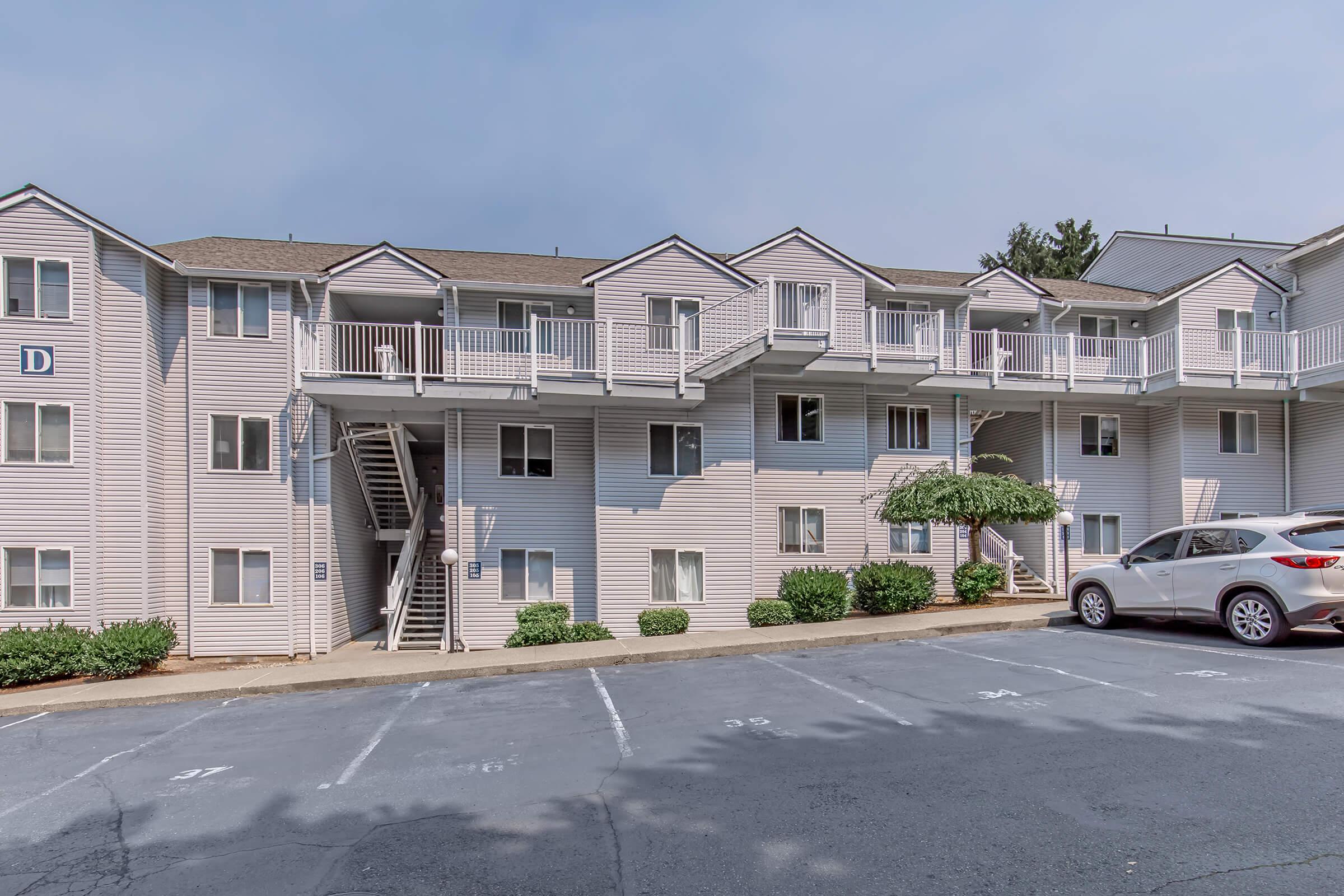 Exterior view of a multi-unit apartment building with light-colored siding. The structure features multiple balconies, greenery around the foundation, and a parking lot with numbered spaces. A white car is parked in front of the building, and the sky is partly cloudy.
