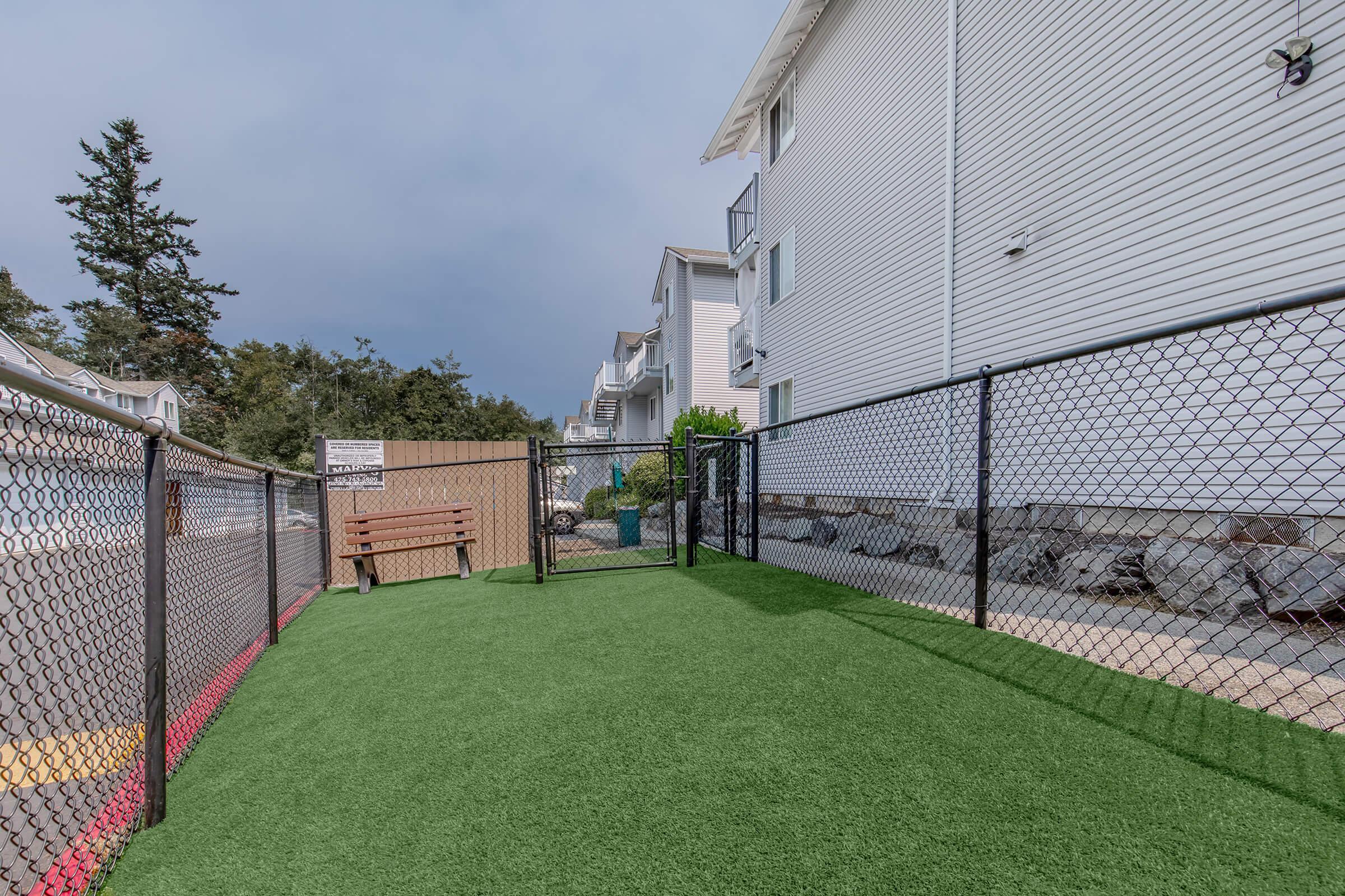 A fenced outdoor area with artificial grass, featuring a bench. Surrounding the space are several apartments, and there are trees in the background. The sky is overcast, adding a gray tone to the scene.
