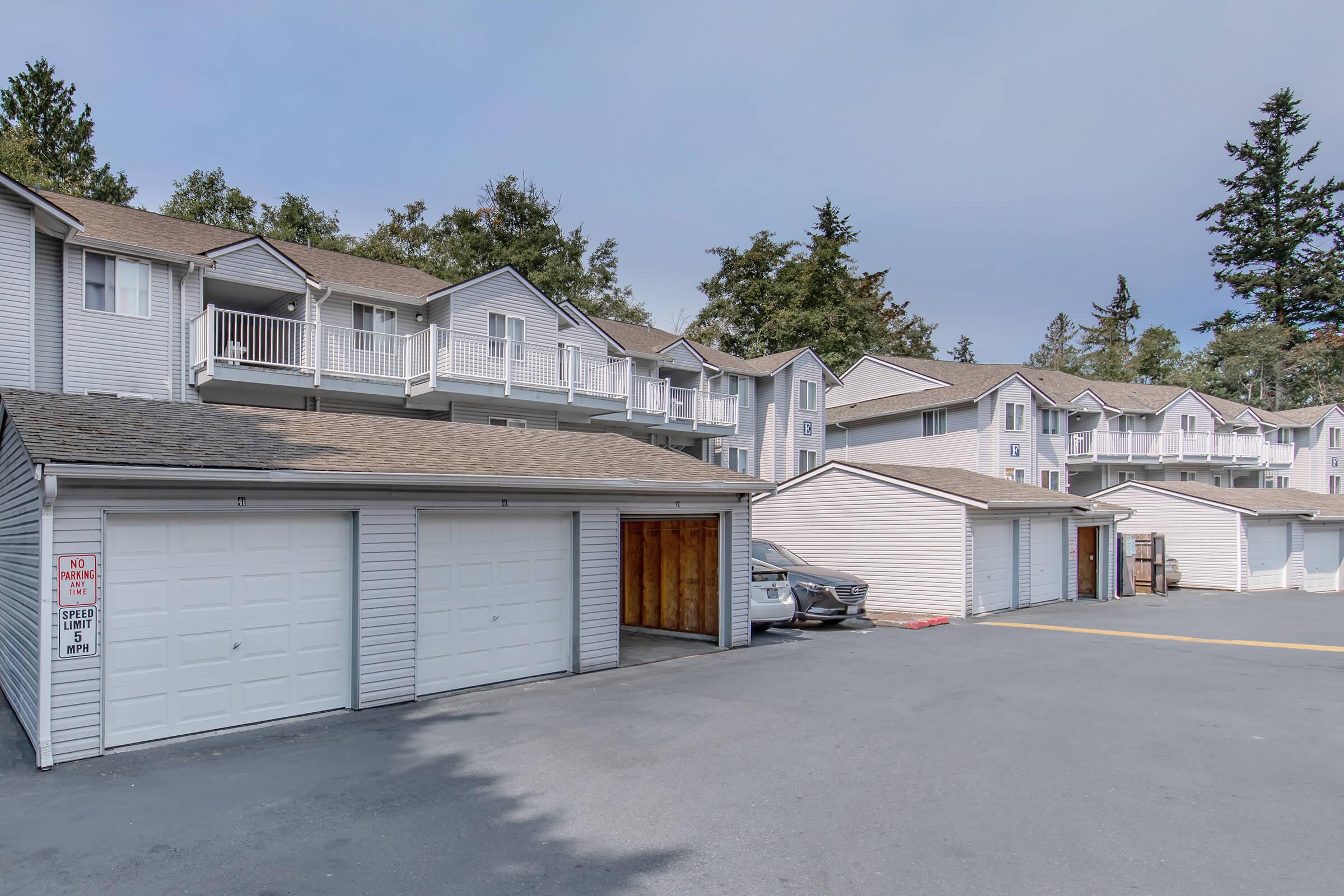 Row of beige and white residential buildings with balconies, featuring multiple garages in the foreground. The scene is set in a quiet area surrounded by trees, and the sky is overcast. Vehicles are parked near the garages, and a designated parking area is visible.