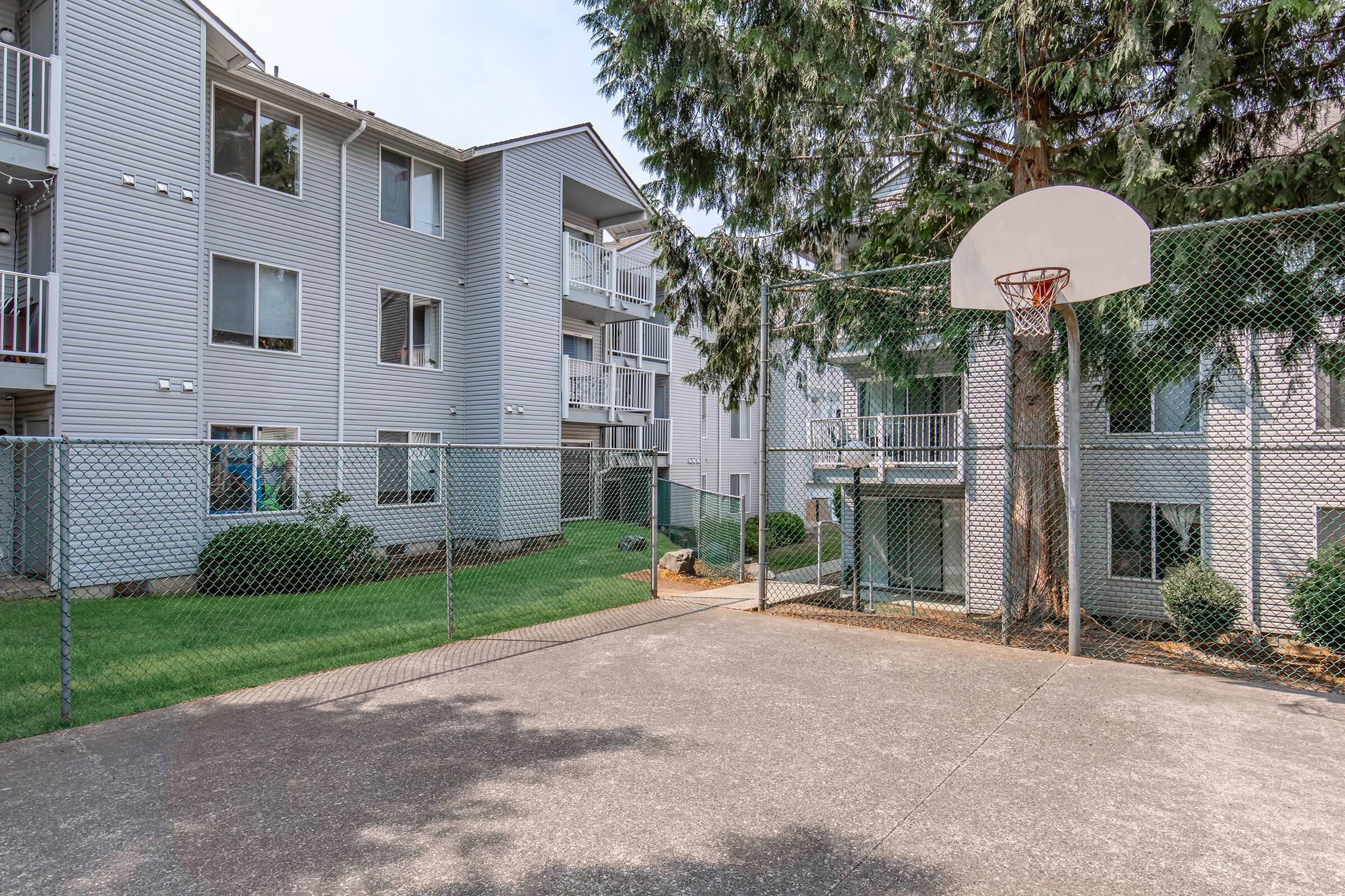 A fenced basketball court with a hoop in the foreground, surrounded by greenery and three-story apartment buildings in the background. The scene is peaceful, with clear skies and well-maintained landscaping.