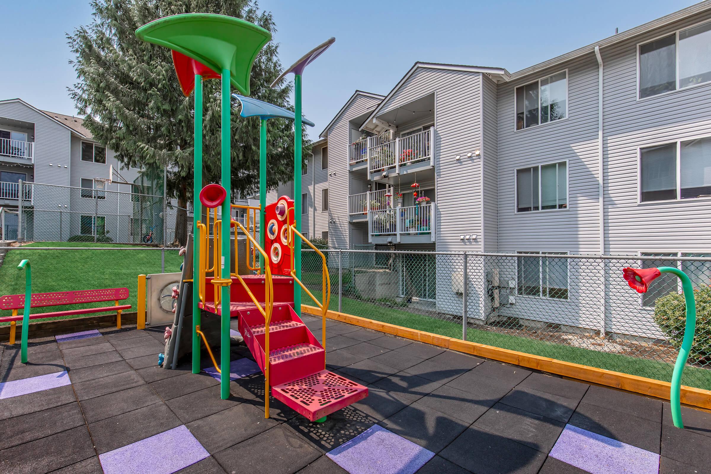 A colorful playground with a play structure featuring slides and climbing structures, surrounded by safety mats. In the background, there are apartment buildings with balconies and a fenced area. The playground displays various tubes and interactive elements for children.