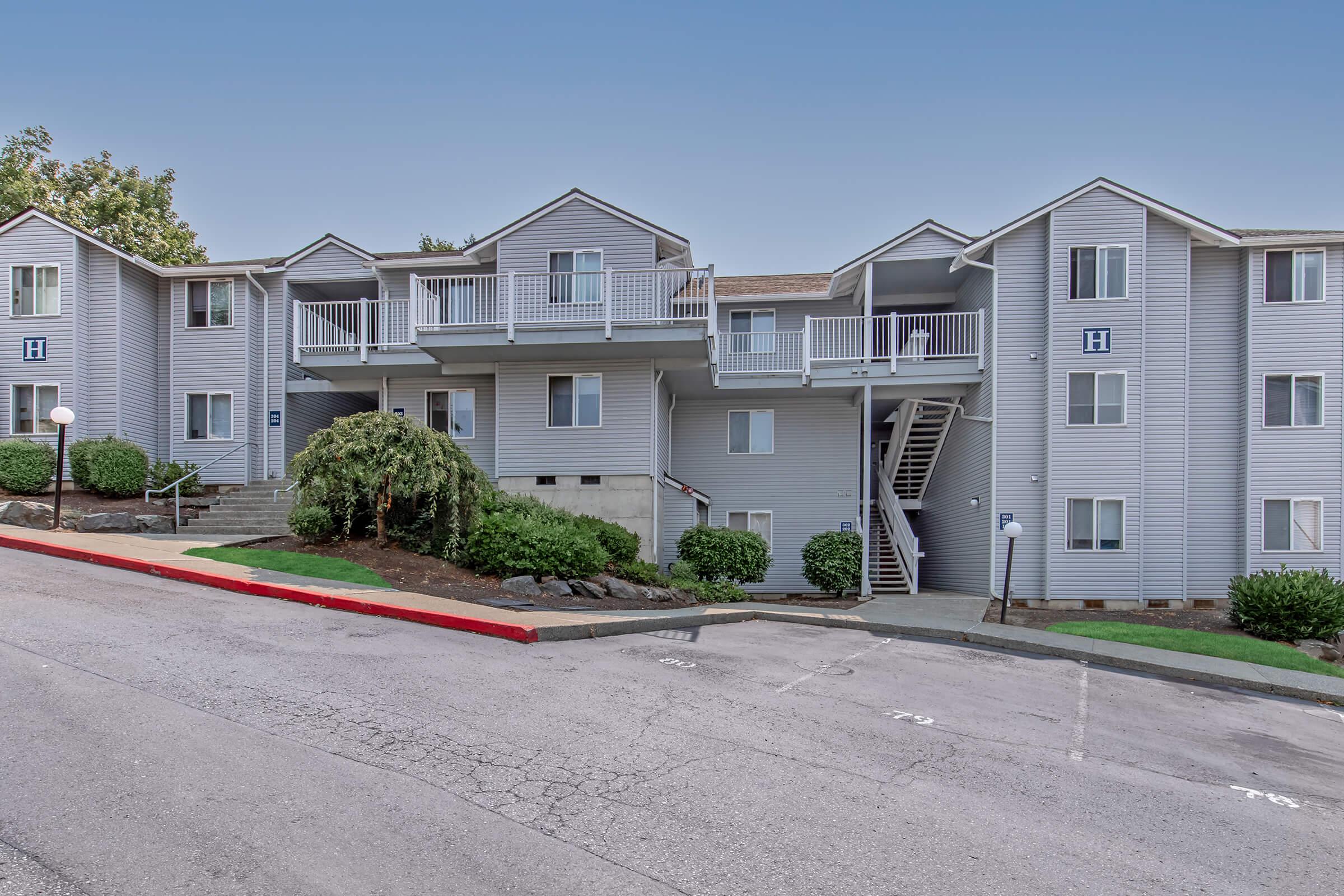 A multi-story apartment building with a light gray exterior. The structure features several balconies and staircases, surrounded by green landscaping and a sloped driveway. The area is well-maintained, with a smooth road leading up to the entrance.
