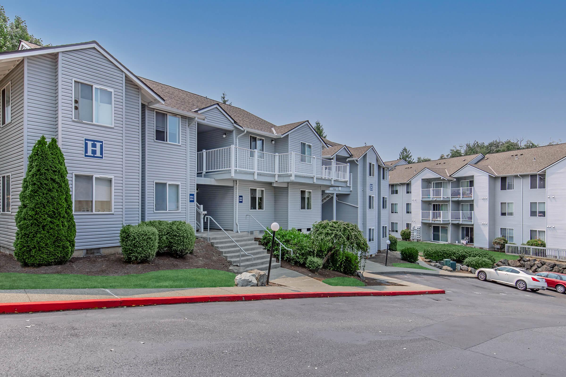 A residential apartment complex featuring multiple two-story buildings with a lightly colored exterior. The scene shows a well-maintained landscape, including shrubs and trees. There are parked cars along the red curb and balconies on the buildings. The sky is clear and blue.