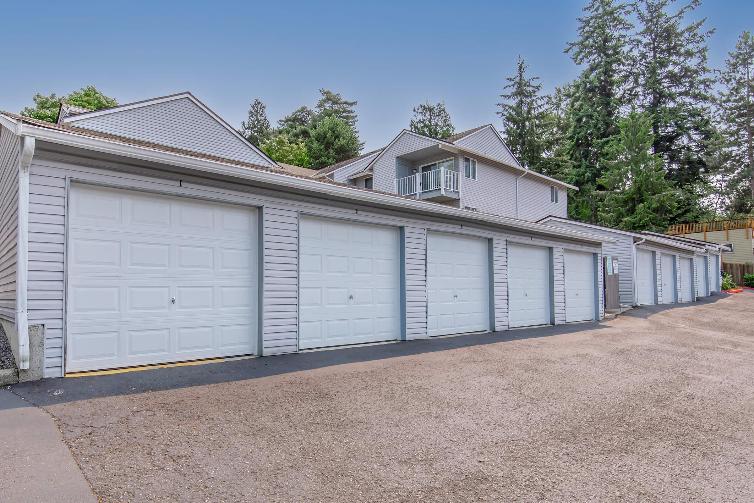 Row of six white garage doors lined up against a light gray building, with trees and greenery in the background. The asphalt driveway leads alongside the garages, and the upper part of the building features a balcony and additional architectural details. Clear blue sky above.