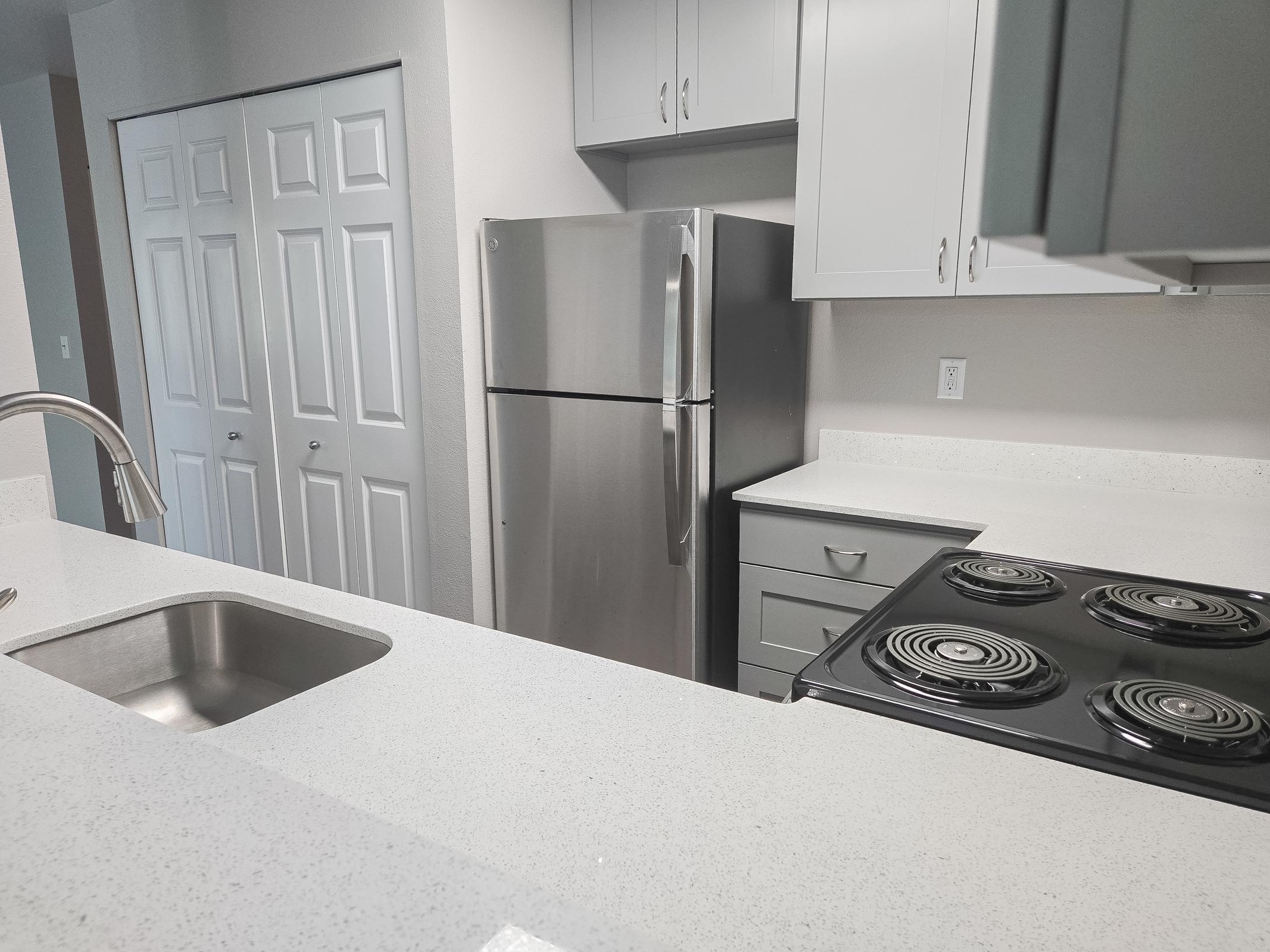 A modern kitchen featuring a stainless steel refrigerator, a white countertop, a black electric stove with spiral burners, and gray cabinets. Closets can be seen in the background, and the overall design is clean and contemporary.