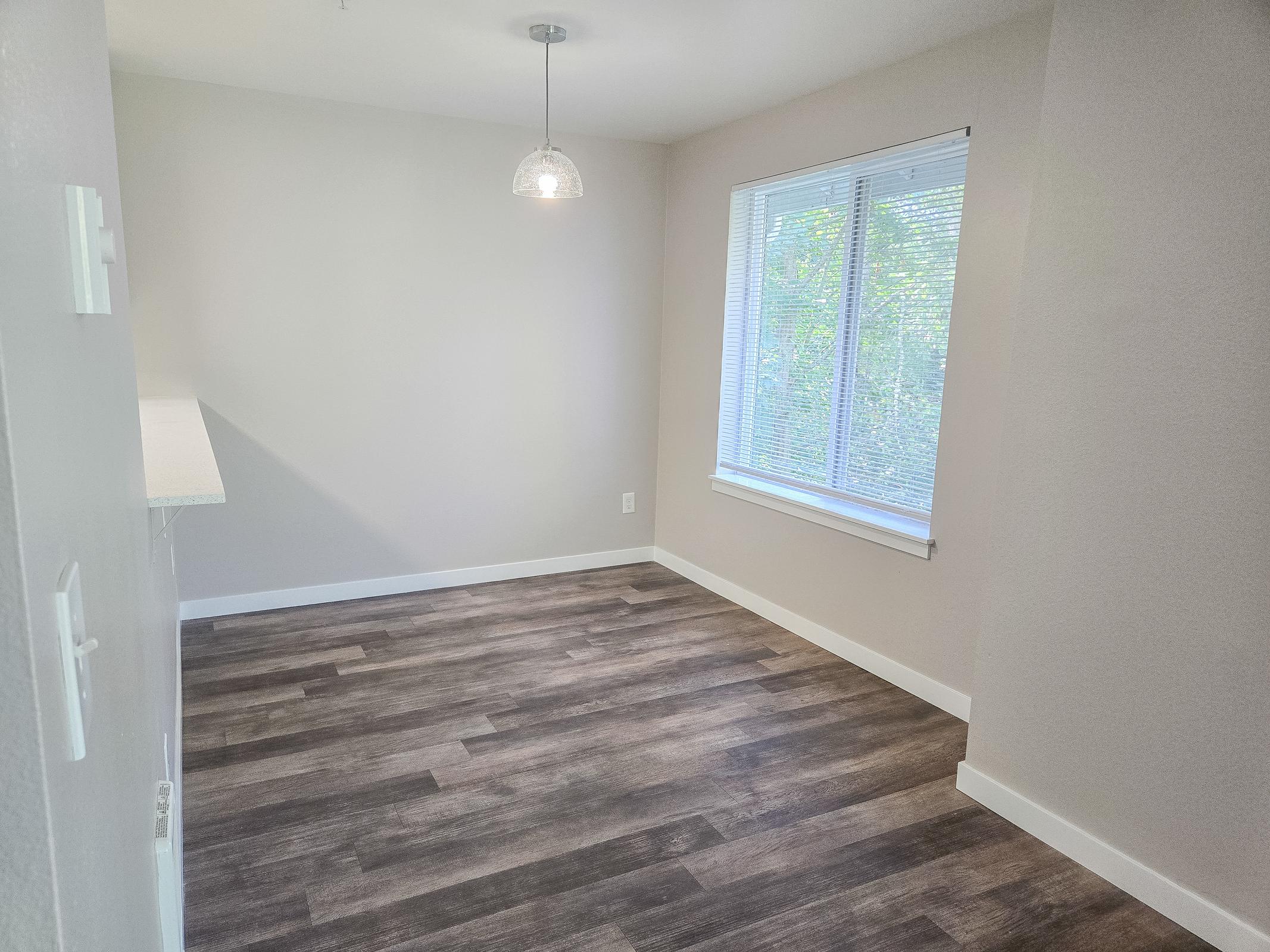 A spacious, empty room featuring light-colored walls, a large window with blinds allowing natural light, and a modern pendant light hanging from the ceiling. The flooring is a dark wood laminate, enhancing the room's clean and open feel.
