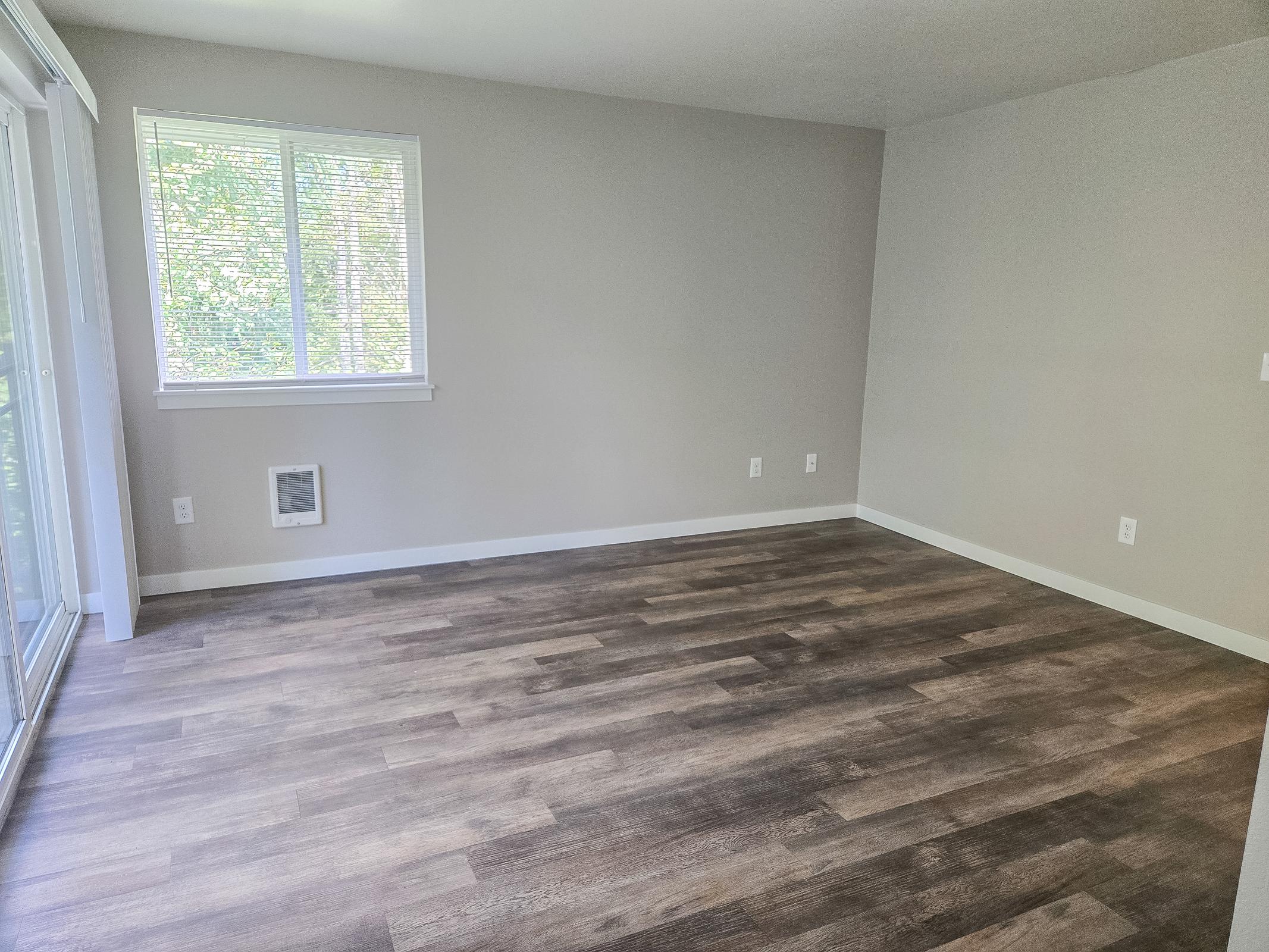 A spacious, empty room with light gray walls and a modern, laminate wood floor. A large window with sheer blinds allows natural light to enter, and there is a small heating vent on one wall. The overall feel is bright and minimalistic, ideal for various uses or customization.