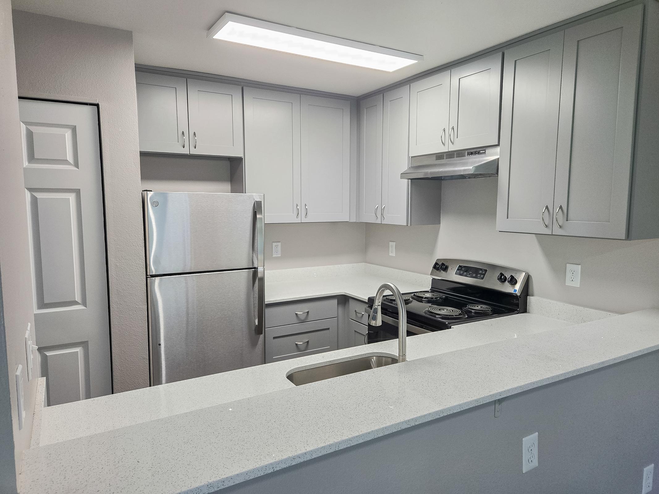 Modern kitchen featuring grey cabinets, a stainless steel refrigerator, a stove with an oven, and a sleek countertop with a sink. Bright lighting enhances the space, which includes a closed door to the left and a clean, minimalist design.