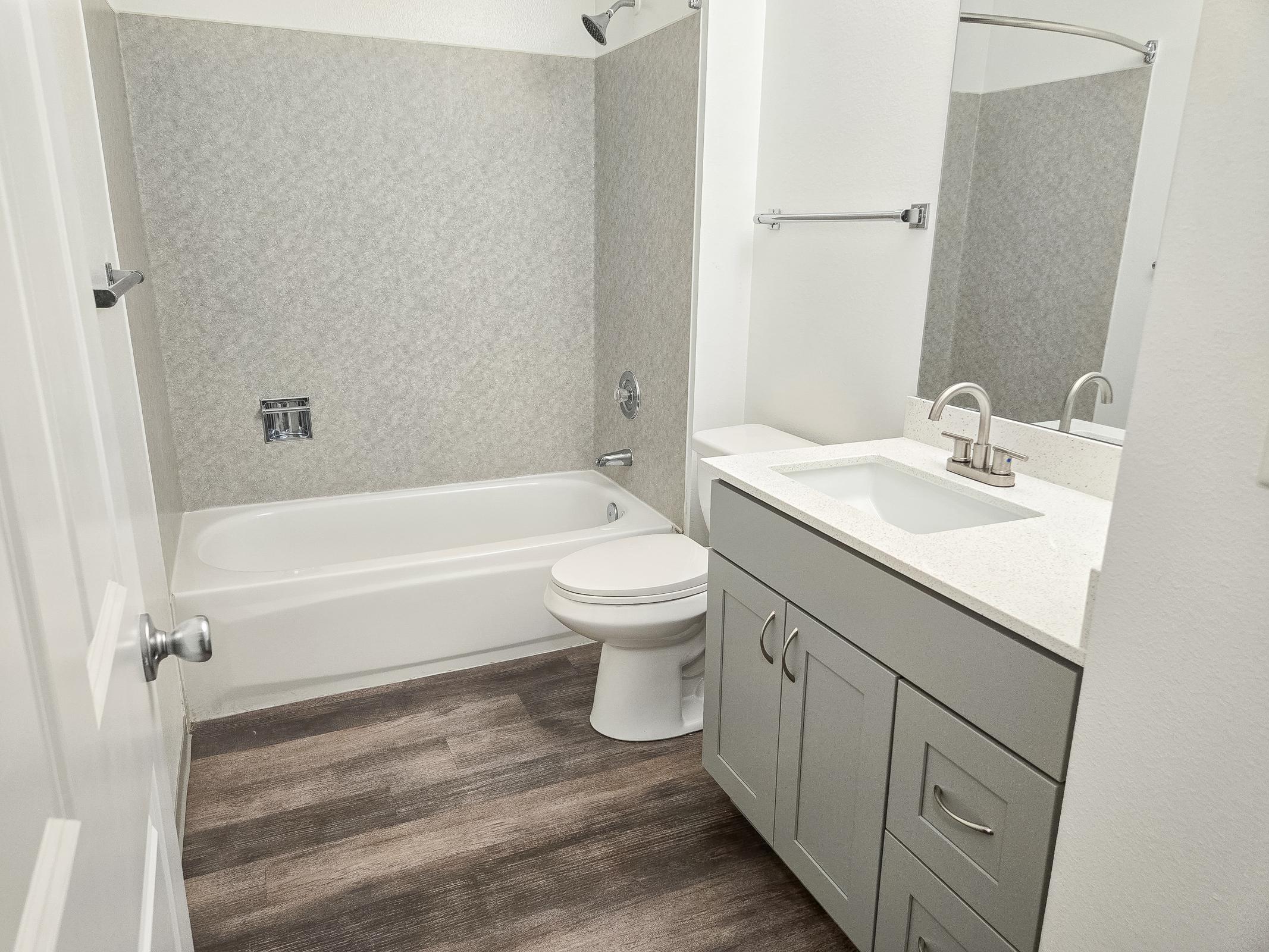 A modern bathroom featuring a bathtub with a shower, a toilet, and a sink with a countertop. The walls are light-colored with patterned accents, and the flooring consists of wooden planks. A mirror is mounted above the sink, and there is a towel bar on the wall.