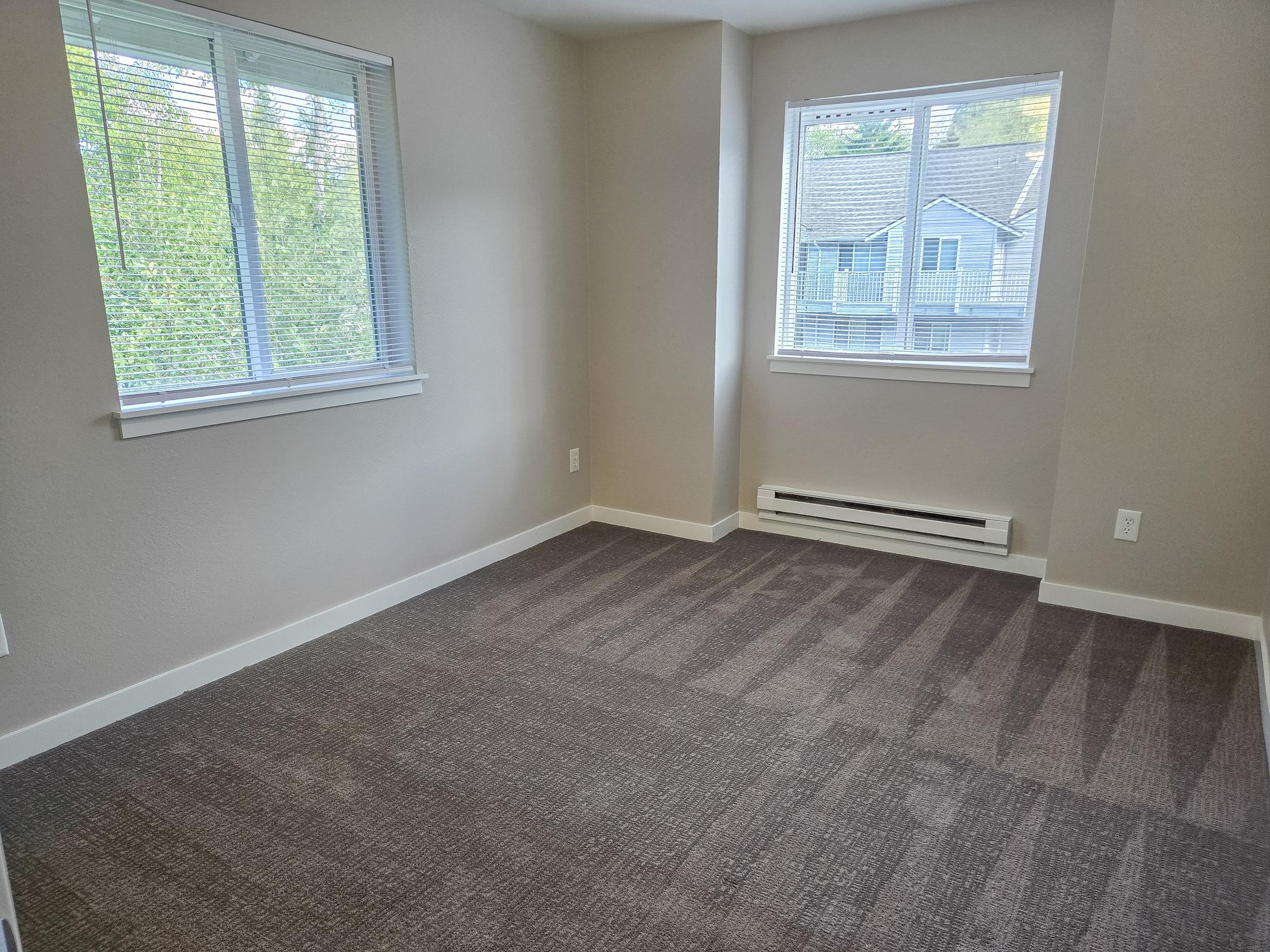 A bright and empty room featuring light gray walls, two windows allowing natural light, and a plush carpeted floor. The space appears clean and freshly painted, with visible baseboards and an air vent along the wall. Ideal for personal customization or as a living space.