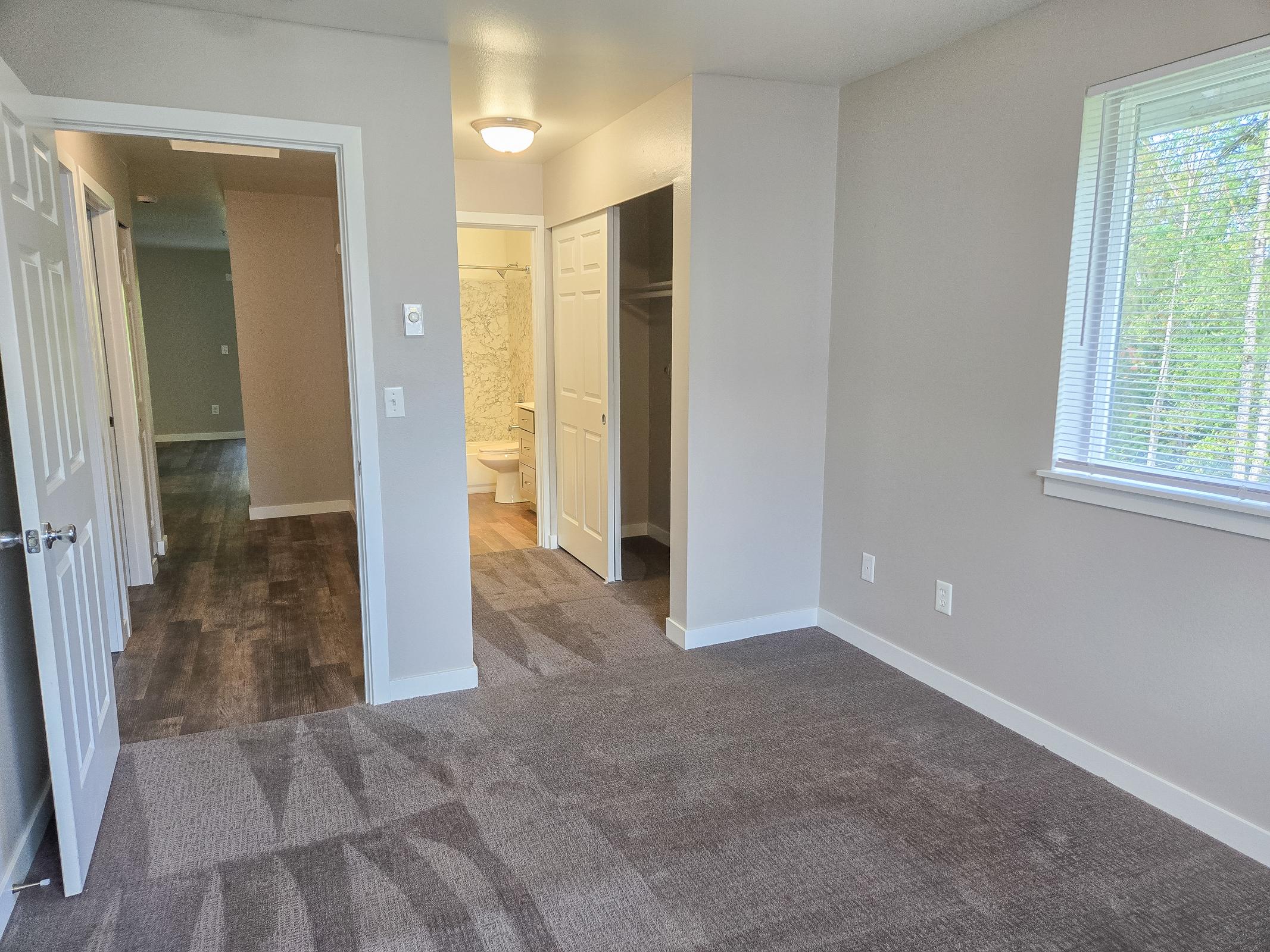 A spacious interior view of a room featuring light gray walls and carpet. The room has an open layout with a doorway leading to a bathroom and closet, alongside a window providing natural light. The overall design is modern and clean, suitable for various residential setups.