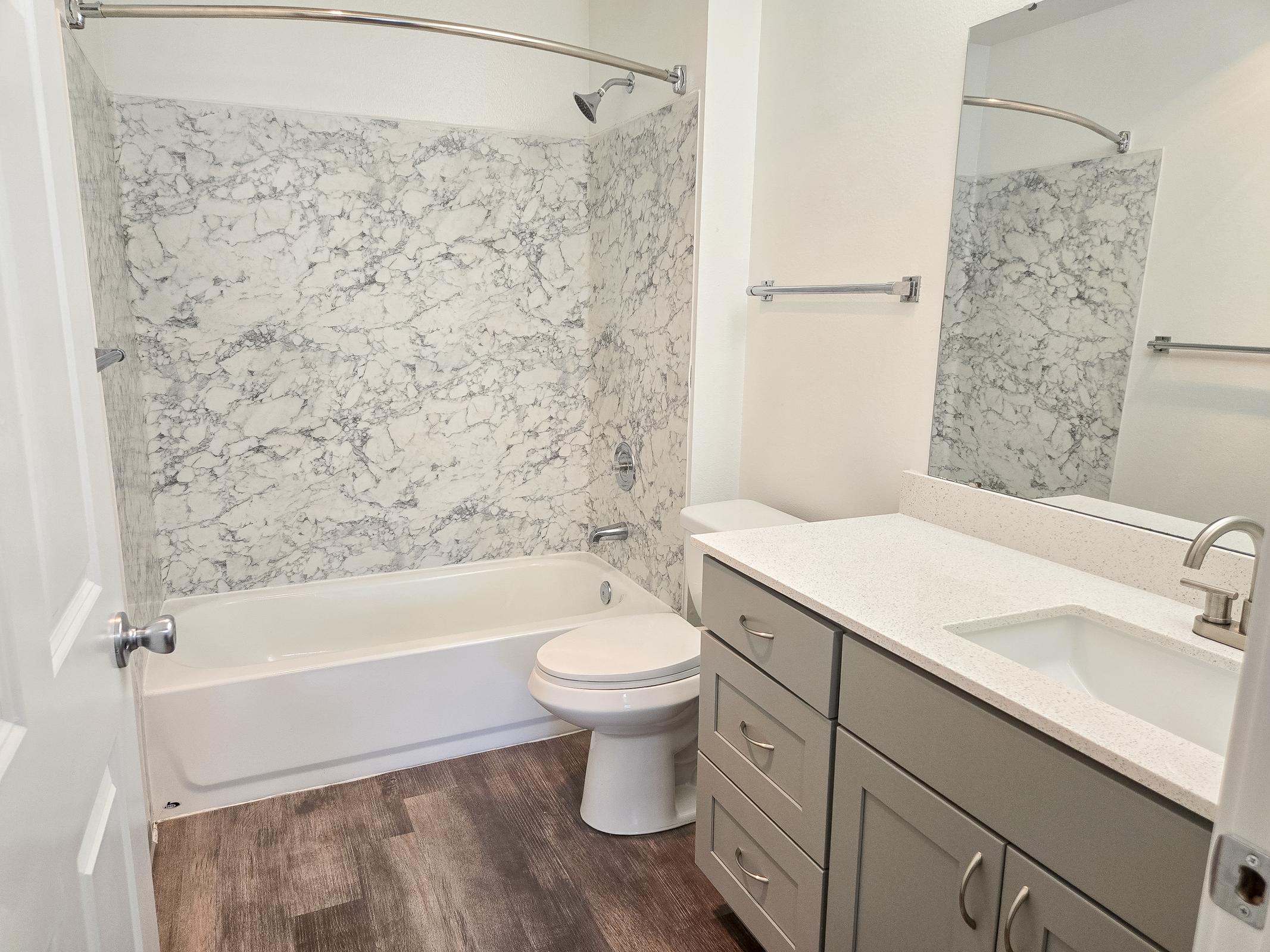 A modern bathroom featuring a white bathtub with a marble-patterned shower wall, a sleek vanity with a sink and gray cabinetry, and a large mirror. The floor has a wood-like finish, providing a warm contrast to the light-colored walls and fixtures.