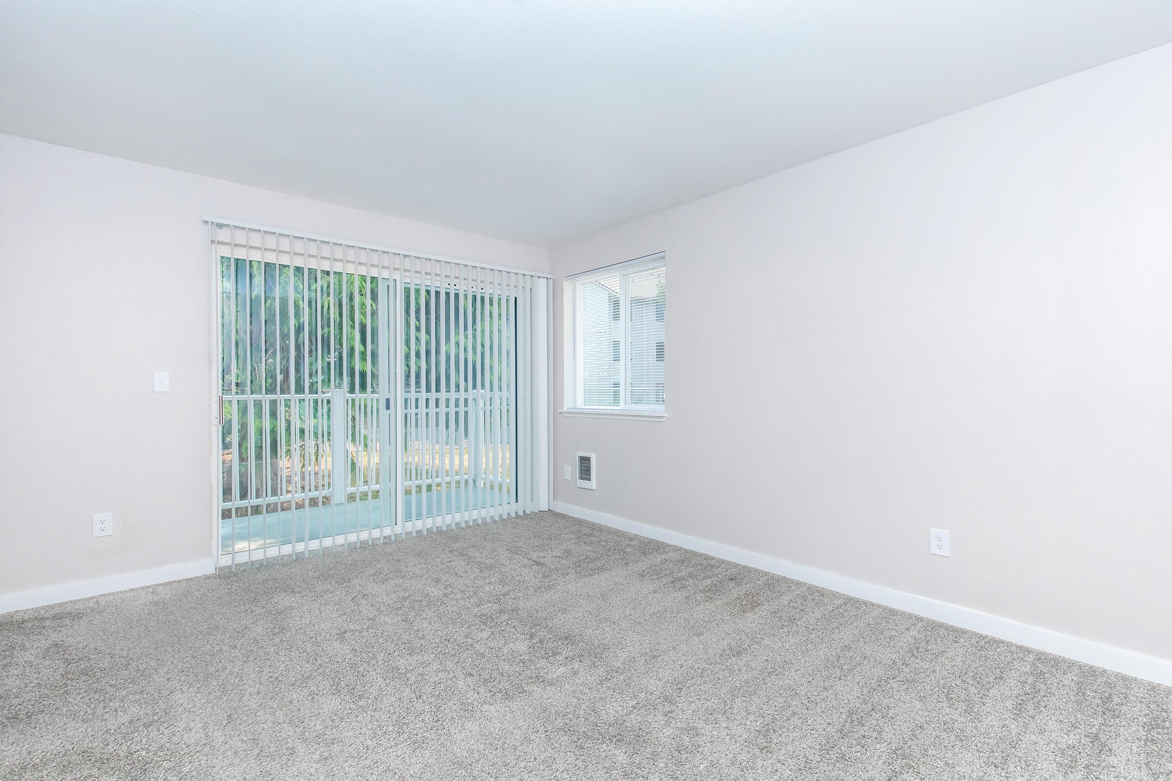 A clean, empty room with light-colored walls and plush gray carpeting. There are large windows allowing natural light, one with vertical blinds leading to an outdoor area. The space appears bright and airy, suitable for various furniture arrangements.