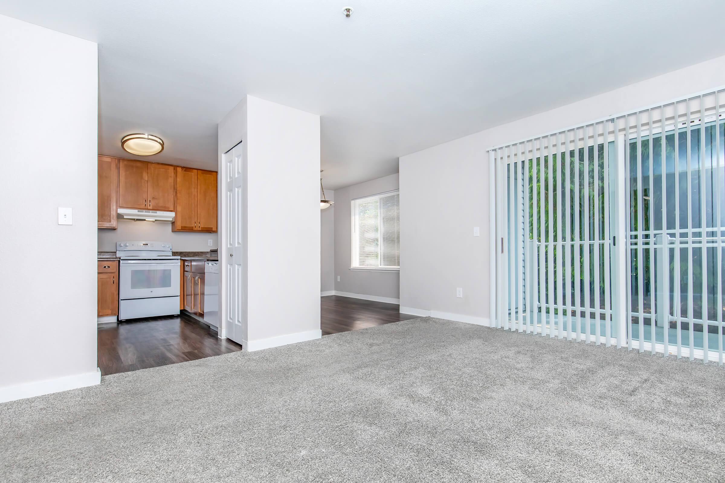 Spacious living area with beige carpet, featuring a doorway leading to a kitchen with wooden cabinets and white appliances. Natural light flows in through large windows and sliding glass doors, which lead to an outdoor space. Simple and modern interior design with neutral colors.