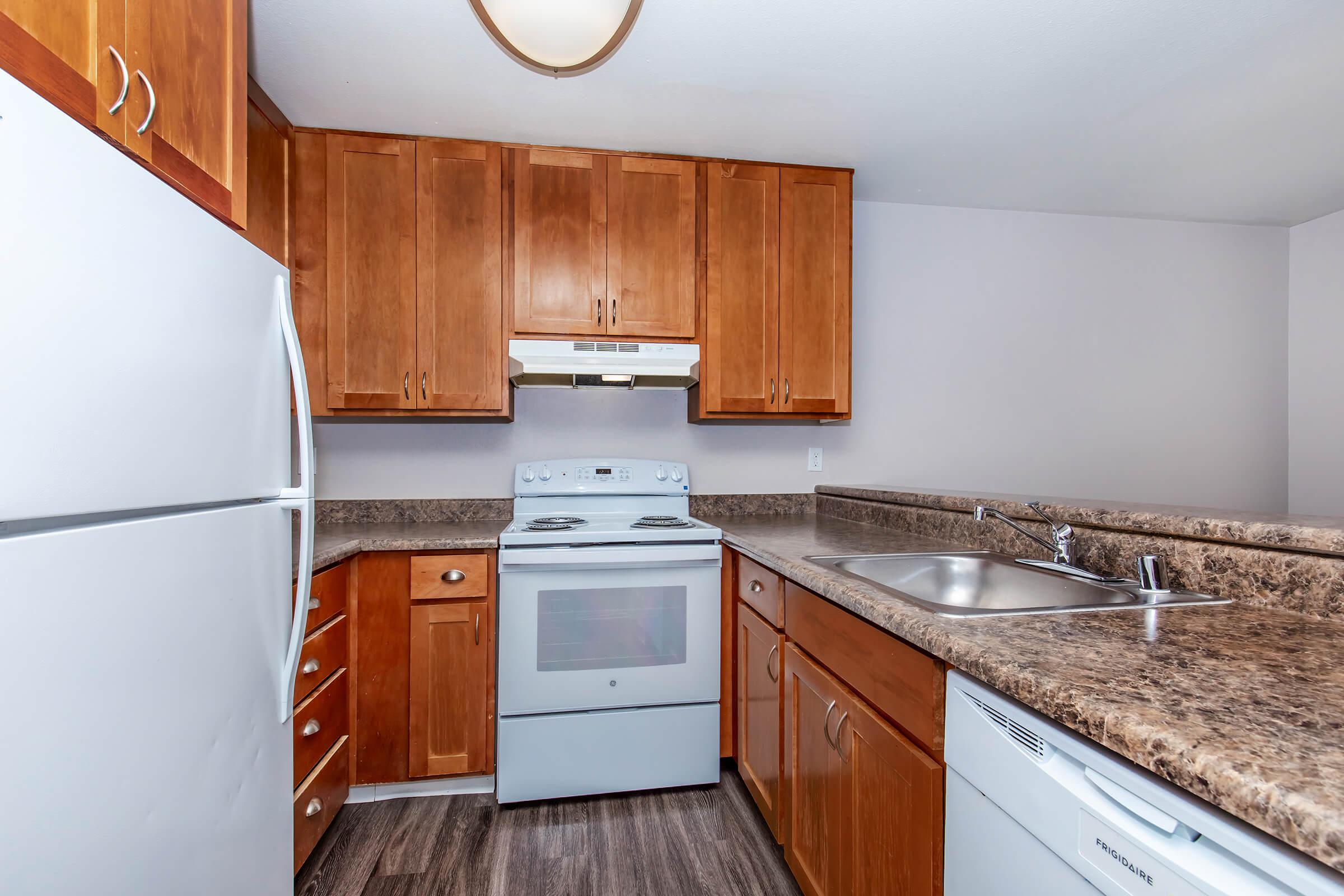 A modern kitchen featuring wooden cabinetry, a white refrigerator, an oven with a range hood, and a stainless steel sink. The countertop is made of a dark, speckled material. The walls are a light color, creating a bright and inviting atmosphere.