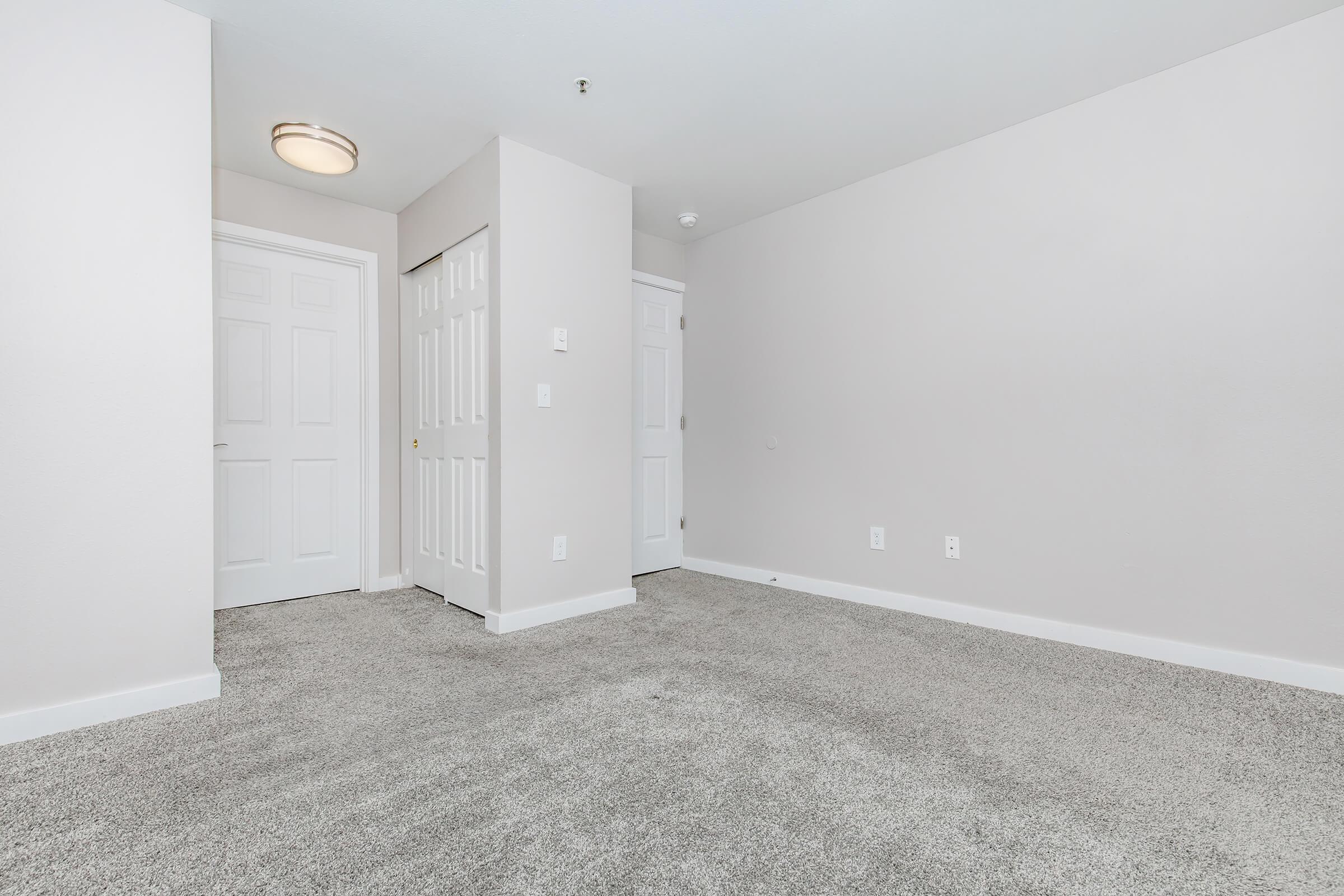 A small, empty room with light gray walls and a carpeted floor. Two doors are visible: one leading to a closet and the other potentially to another room or hallway. A ceiling light is mounted above, adding bright but soft illumination to the space. The overall look is clean and minimalistic.