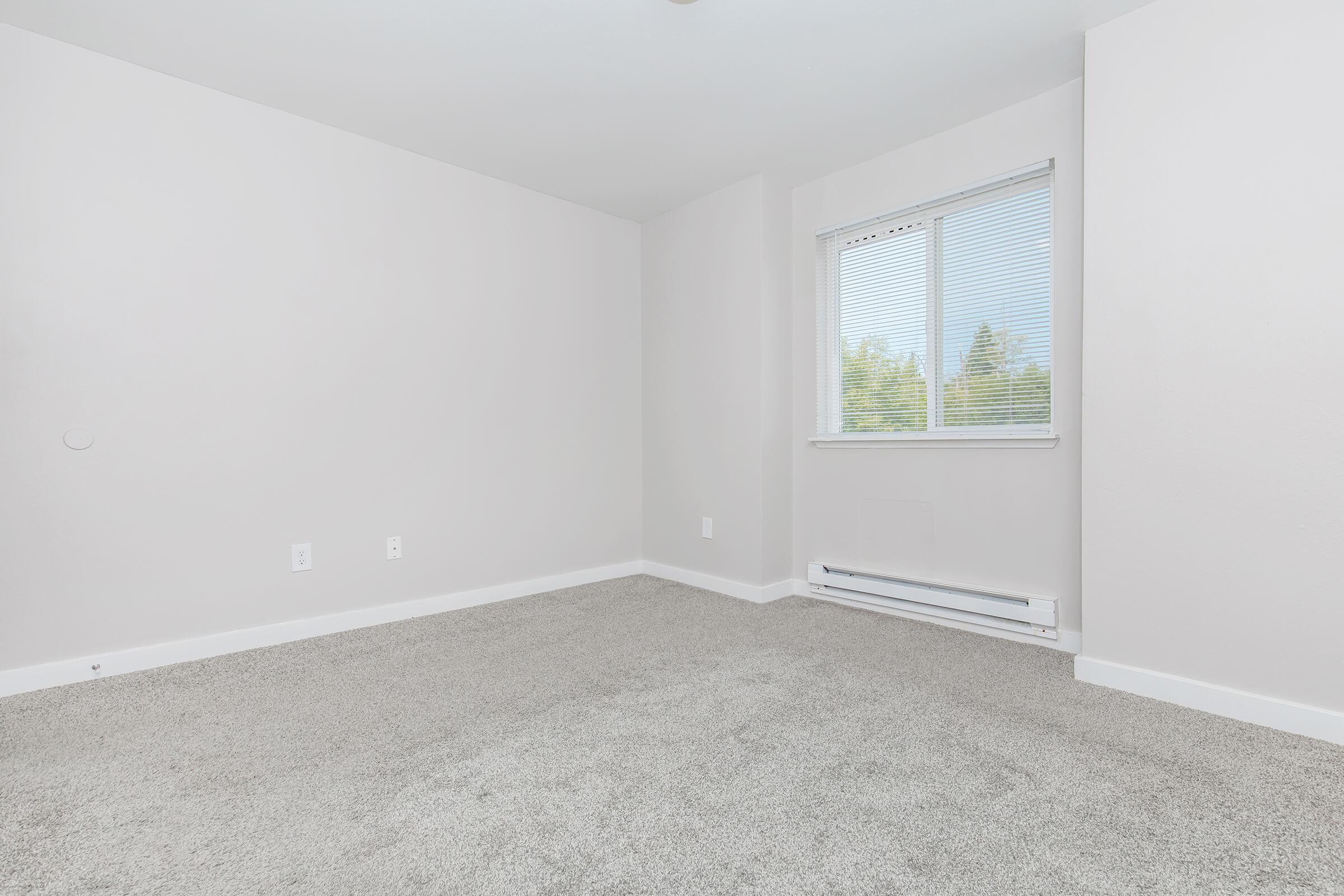 A spacious, empty room with beige walls and light gray carpet. A small window with white blinds allows natural light to enter, illuminating the area. The room has no furniture, creating a clean and minimalistic atmosphere.