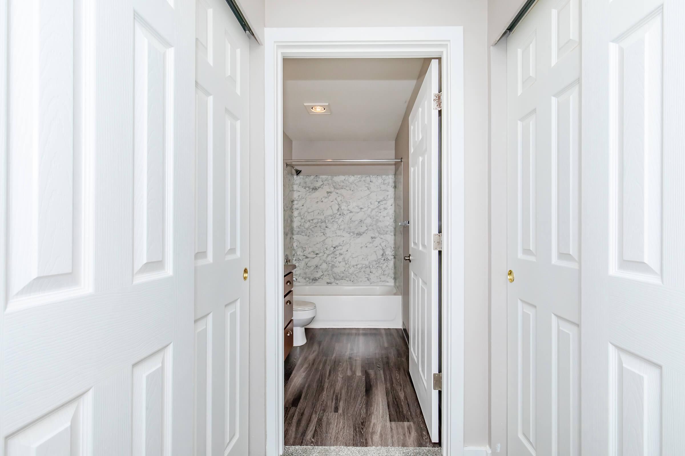 View of a hallway leading to a bathroom, featuring two closed white doors. The bathroom has a marble wall behind a bathtub, wooden cabinetry, and a neutral color scheme. The flooring is a dark wood, complementing the modern design of the space. Natural light enters the area from above.