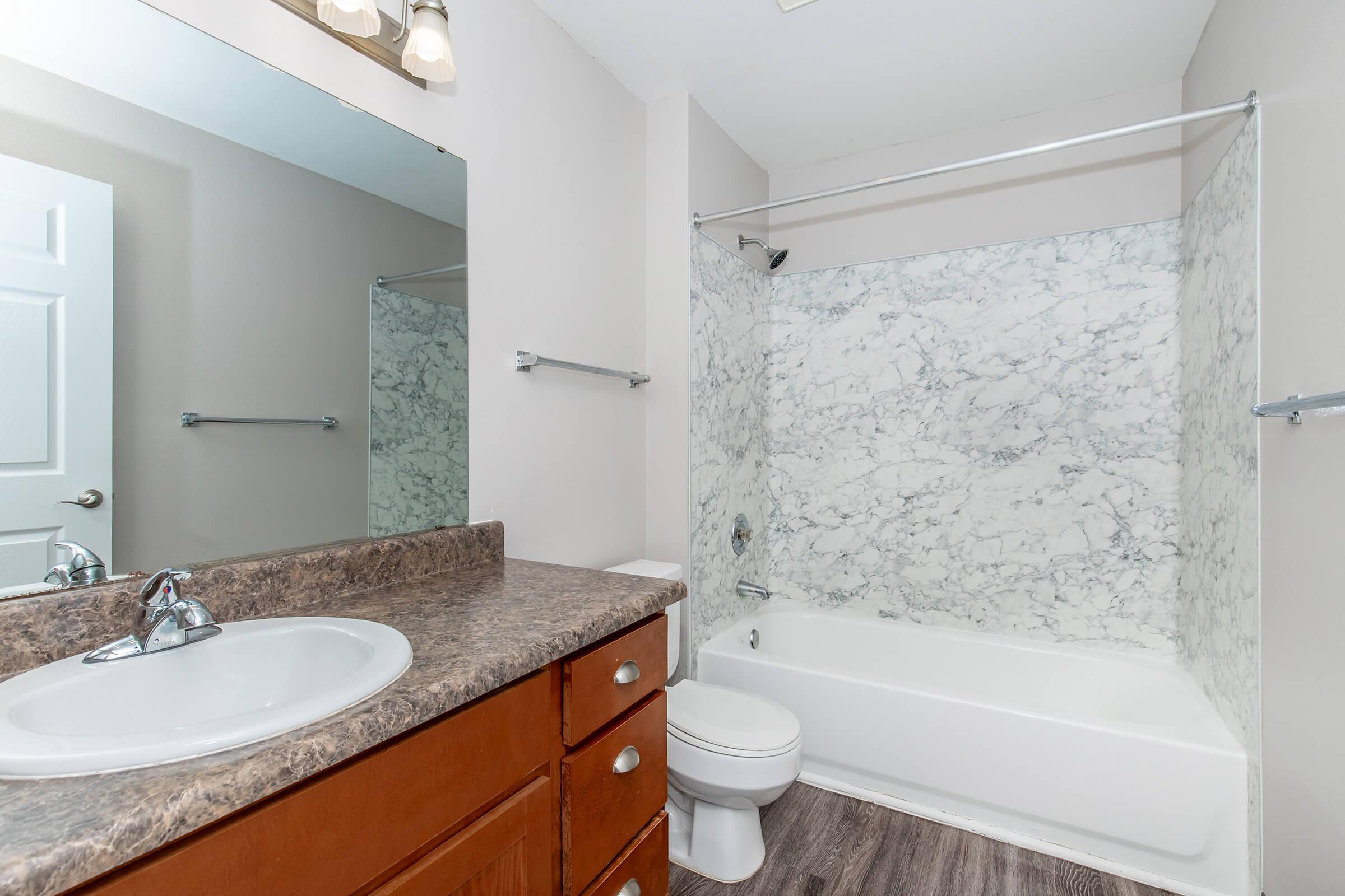 A clean and modern bathroom featuring a bathtub with a marble-patterned shower wall, a sink with a granite countertop, and a white toilet. The room has neutral-colored walls, a large mirror above the sink, and wooden cabinetry for storage. Bright lighting enhances the spacious feel of the area.