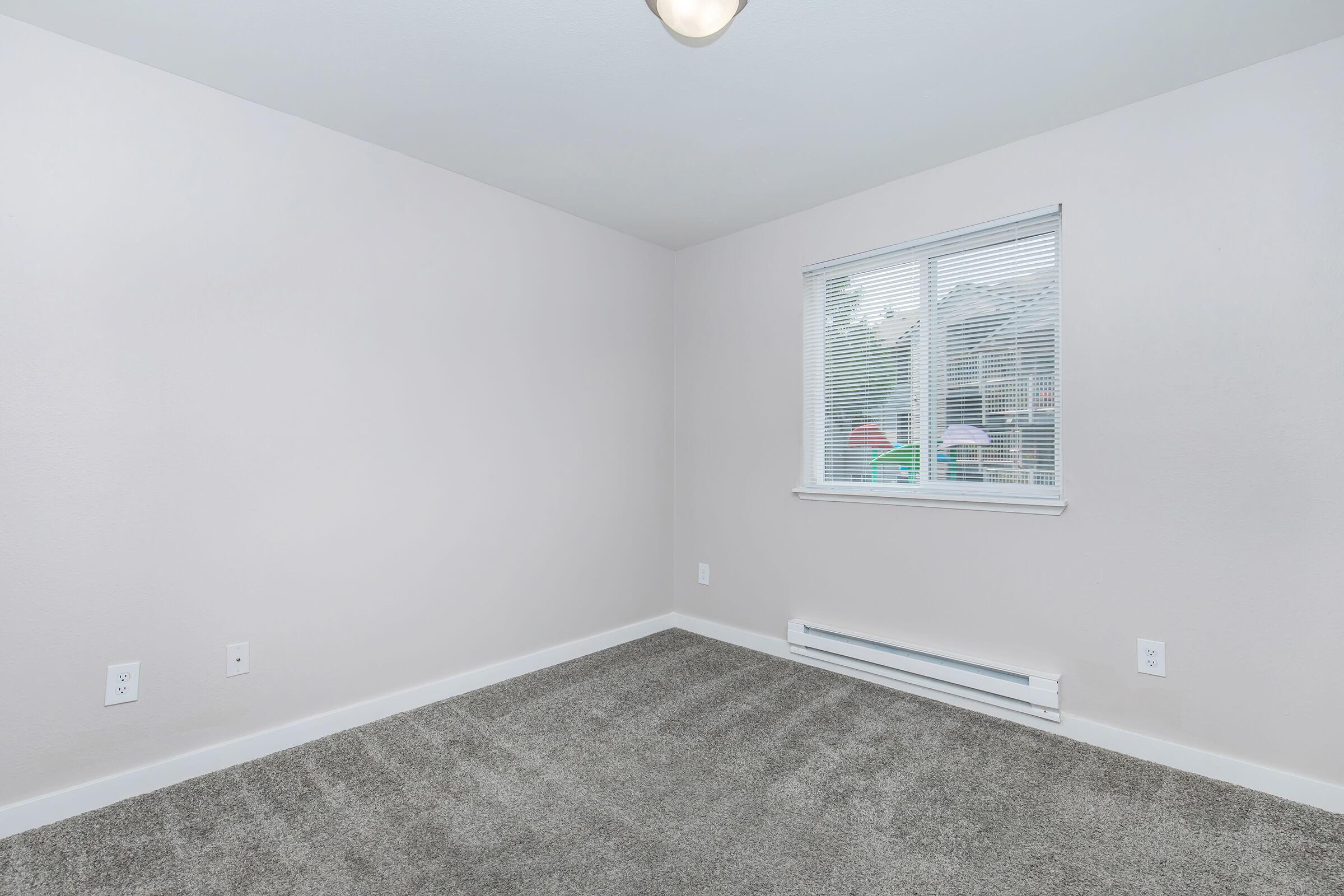 A vacant room with light gray walls and a carpeted floor. There is a window with blinds allowing natural light to enter, and baseboard heating visible below the window. The room appears clean and freshly painted, with no furniture or decorations.