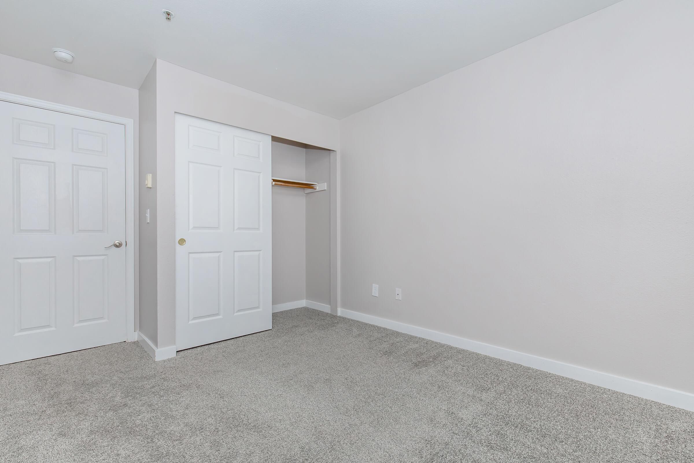 A clean, empty room with light gray walls and carpeted flooring. There is a closet with double doors on the left, which is open and has a wooden shelf inside. The room features no furniture, allowing for a spacious feel.