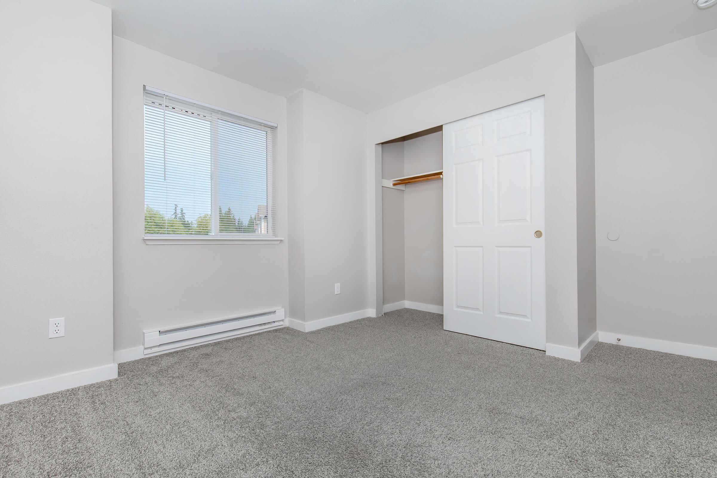Empty room with light gray walls and carpet. Features a window with blinds allowing natural light, a closet with a white door, and a heating vent along the wall. The room is spacious and uncluttered, creating a clean and fresh appearance.