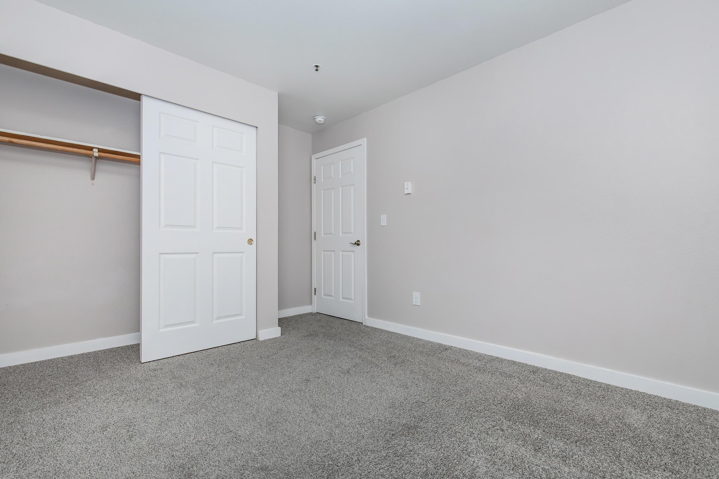 A light grey bedroom with freshly painted walls and carpeted floor. There is a closet with a wooden shelf and a closed door. The opposite wall features a white door leading to another room, creating a simple and clean layout. No furniture or decorations are present in the space.