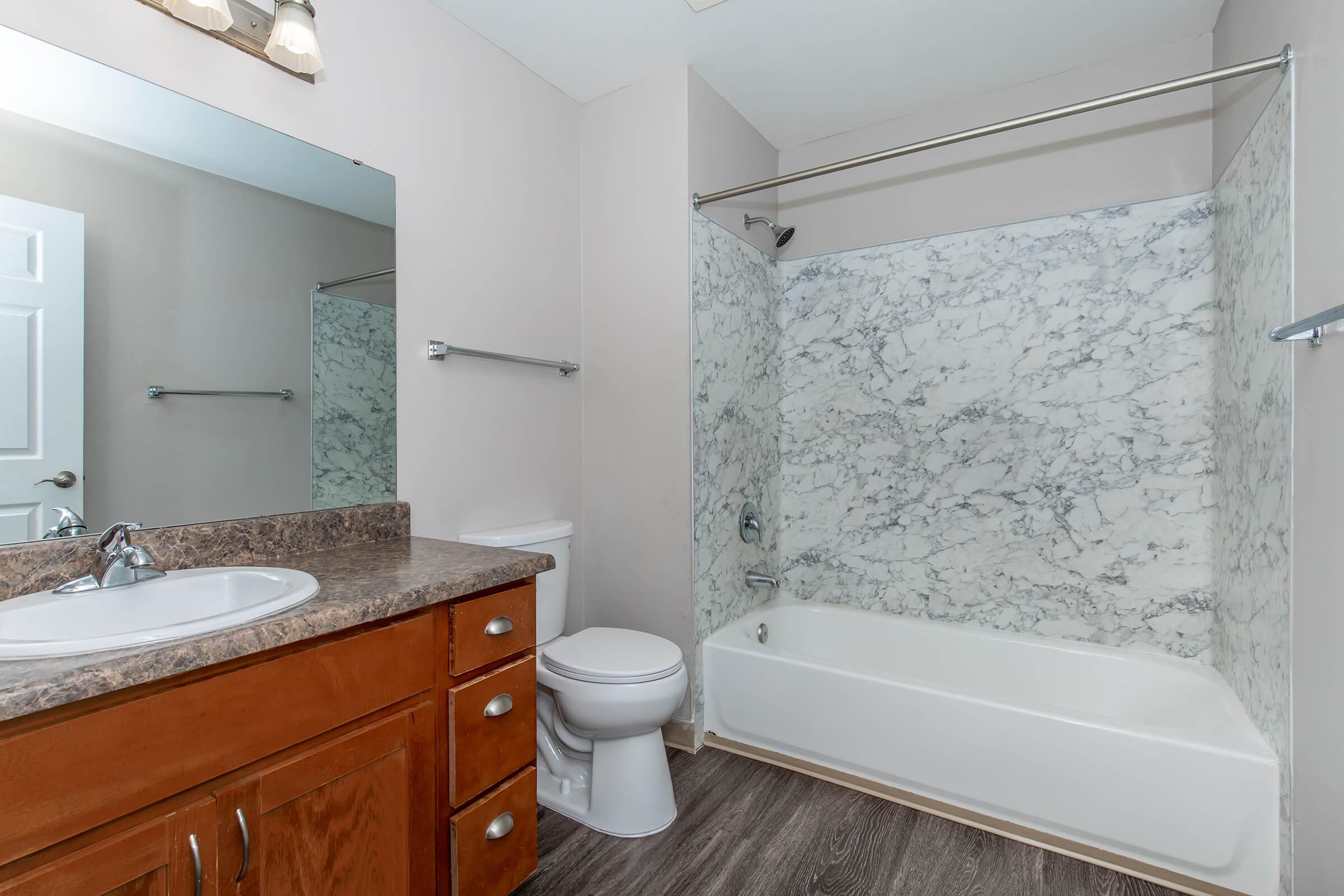 A modern bathroom featuring a white bathtub with a shower, marble-patterned wall tiles, a wooden vanity with a sink, a large mirror, and a toilet. The room has neutral-colored walls and a wooden floor, creating a clean and contemporary look.