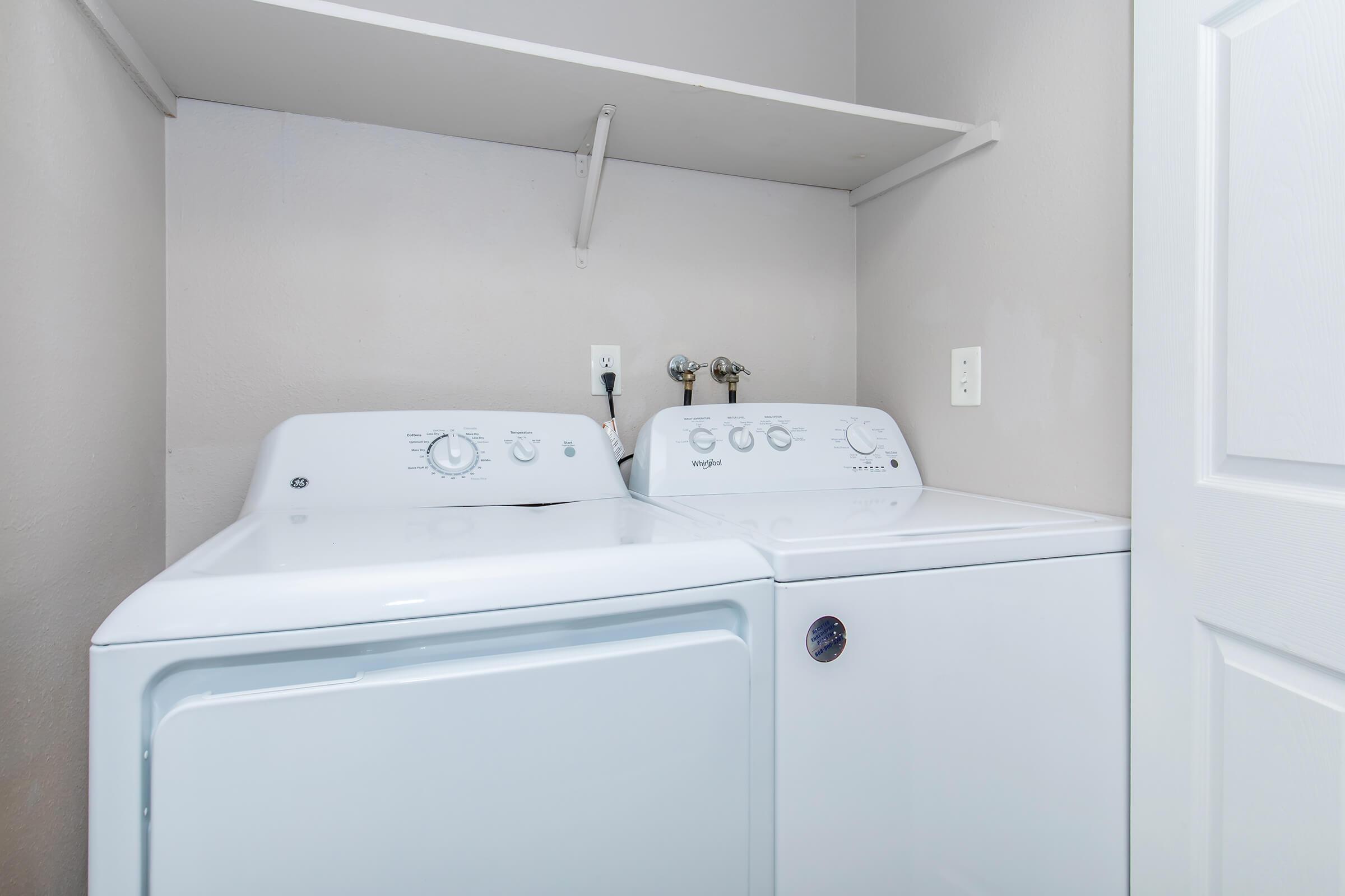 A compact laundry room featuring a white washing machine and dryer side by side. The machines have simple control dials and are set against a light gray wall, with a shelf above them for storage. A door is partially visible on the right.
