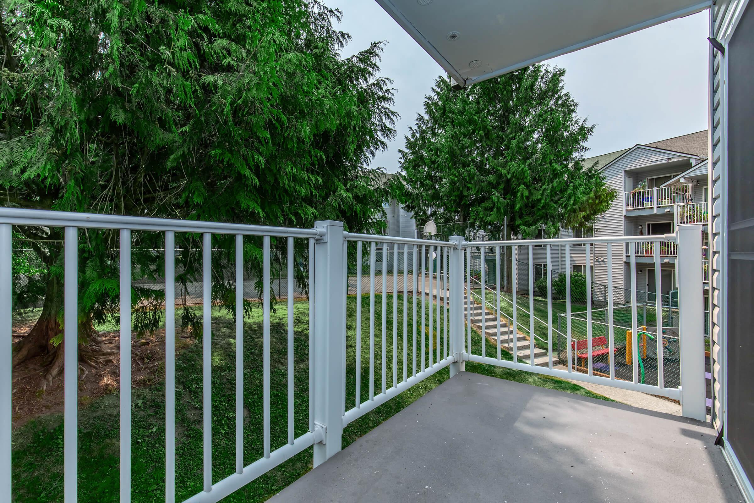 A balcony view featuring white railing overlooking a lush, green landscape with trees. In the background, there are apartment buildings and a playground visible. The scene is bright and peaceful, conveying a serene outdoor environment.