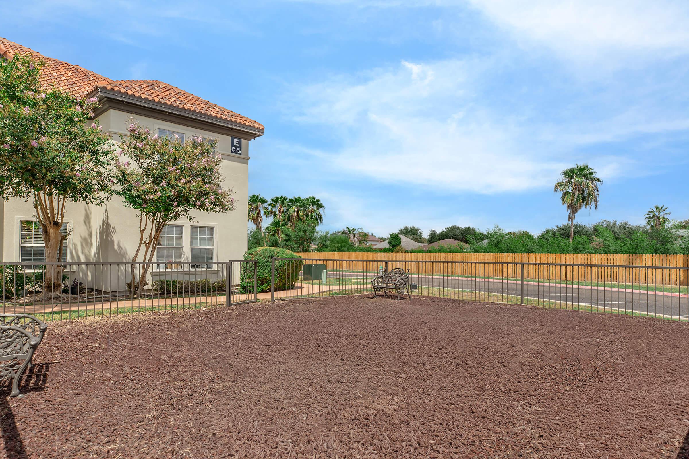 A landscaped outdoor area featuring a fenced-in space with brown wood chips, two benches, and a view of palm trees in the background. A building with a red-tiled roof is to the left, and a fence marks the perimeter of the property. The sky is clear with a few clouds.
