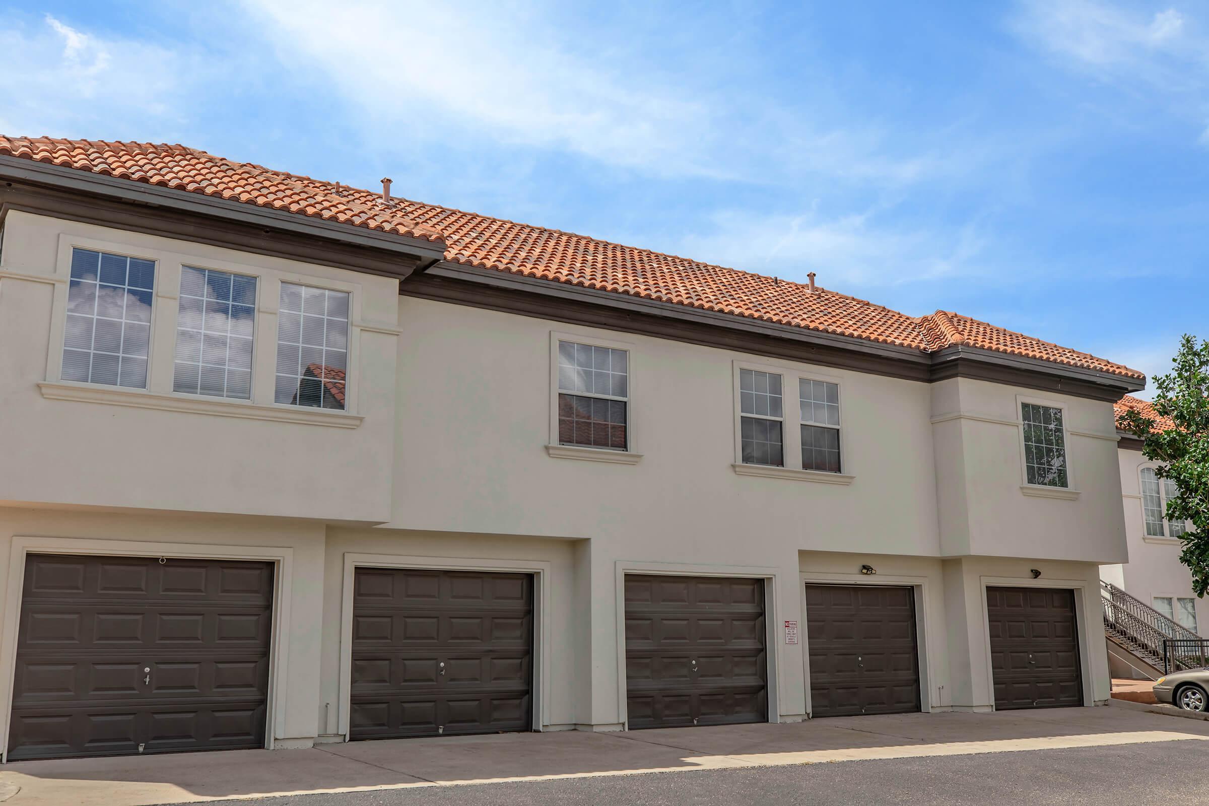 A row of two-story apartment buildings with brown garage doors. The buildings feature large windows and terracotta tiled roofs, set against a clear blue sky. A small tree is visible near the sidewalk in front.