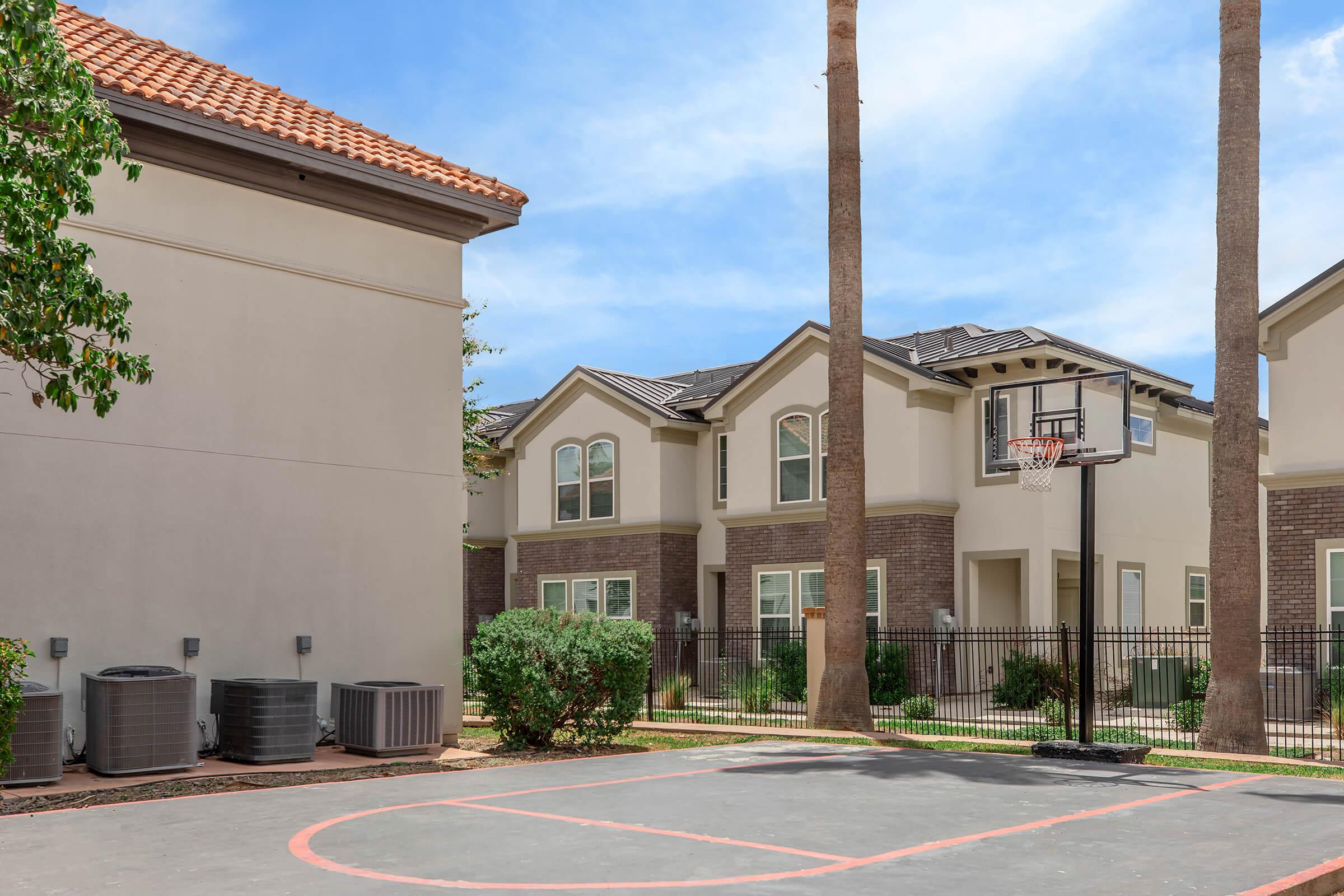 View of a residential area featuring a basketball court surrounded by palm trees and two-story apartment buildings. The buildings have a modern design with brick accents and large windows. Air conditioning units are visible near the court, indicating a warm climate.