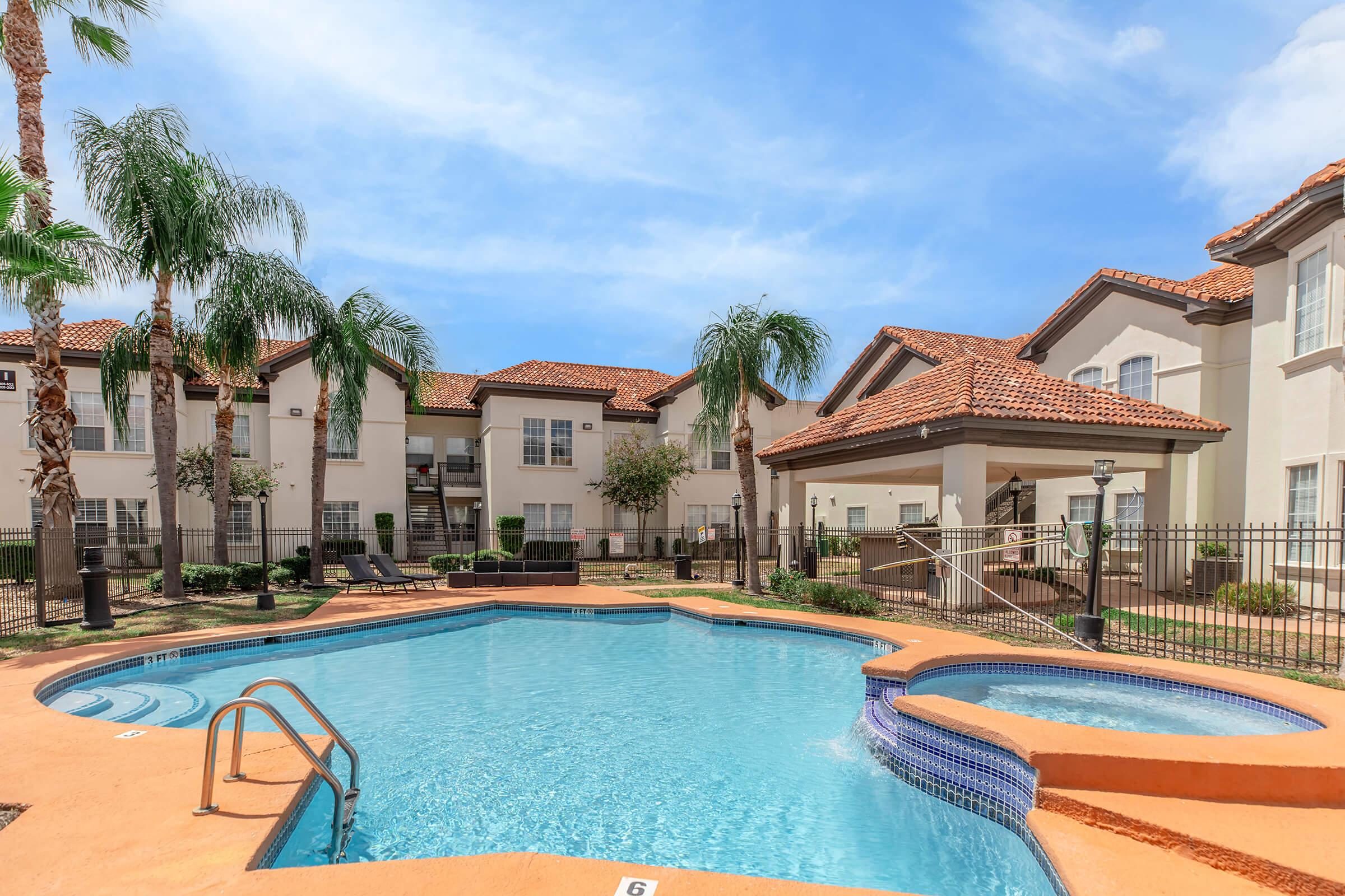 A swimming pool surrounded by palm trees, lounge chairs, and apartment buildings with a Mediterranean-style architecture. The pool area features a unique shape with a shallow spot, and the sky is clear with a few clouds.