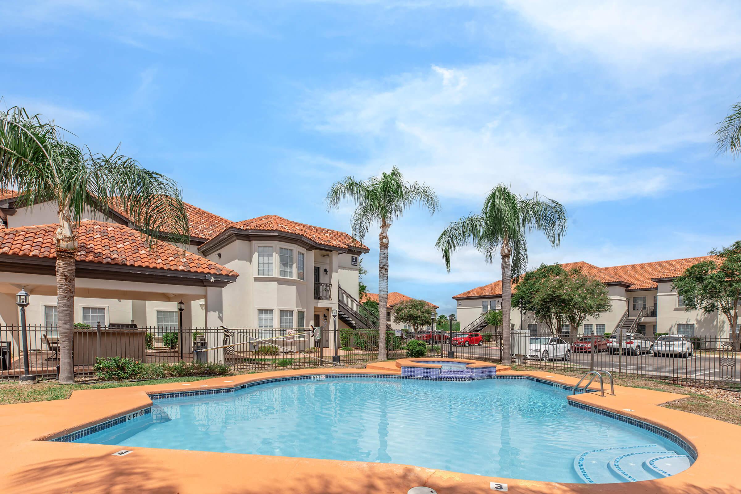 A tranquil residential area featuring a swimming pool surrounded by palm trees and well-maintained landscaping. In the background, there are multi-story apartment buildings with a red tile roof under a clear blue sky. The area is designed for relaxation and leisure.