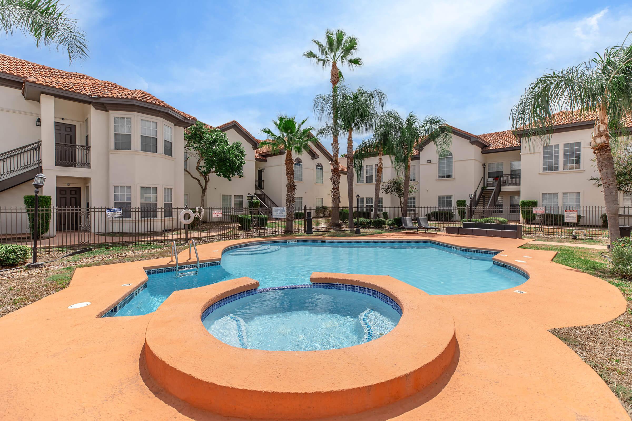 A bright outdoor swimming pool surrounded by palm trees and landscaped greenery, featuring a separate hot tub area. The background shows apartment buildings with balconies and a clear blue sky. Sun loungers are placed around the pool area, creating a relaxing atmosphere.