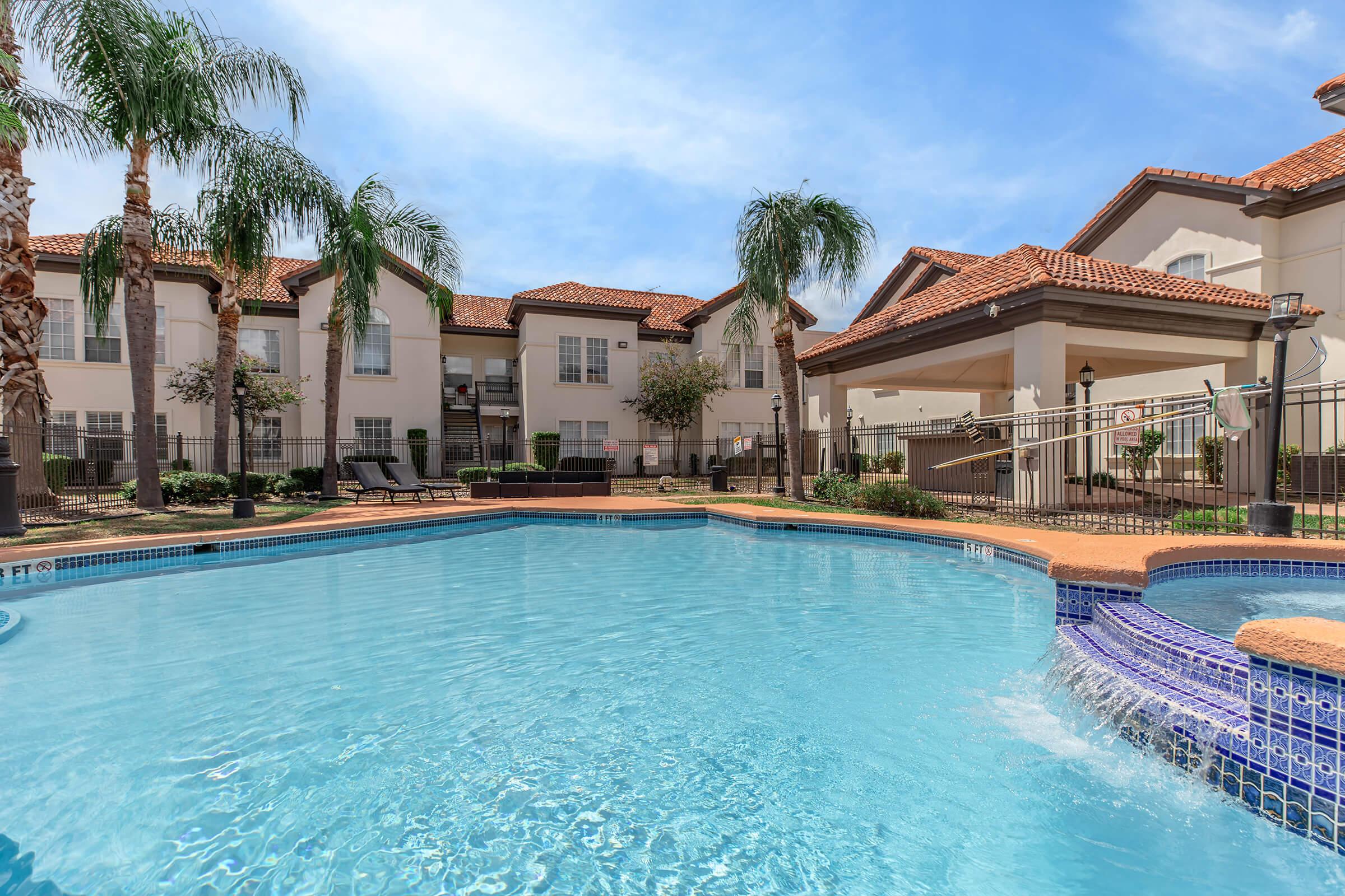 A bright blue swimming pool surrounded by palm trees and landscaped gardens, with several lounge chairs nearby. In the background, residential buildings with orange tile roofs can be seen under a clear blue sky. The pool area is fenced and well-maintained, creating a relaxing atmosphere.