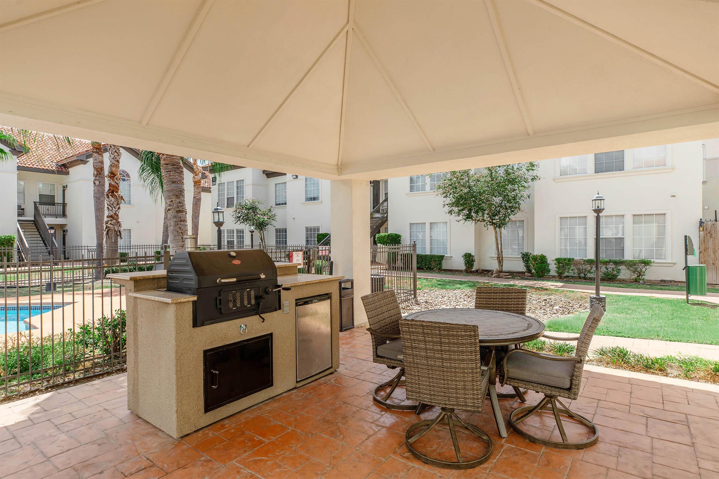 Outdoor barbecue area featuring a grill, a round table with four chairs, and a shaded gazebo. The background includes a well-maintained lawn with shrubs and palm trees, along with apartment buildings visible in the distance. The setting is ideal for outdoor dining and social gatherings.