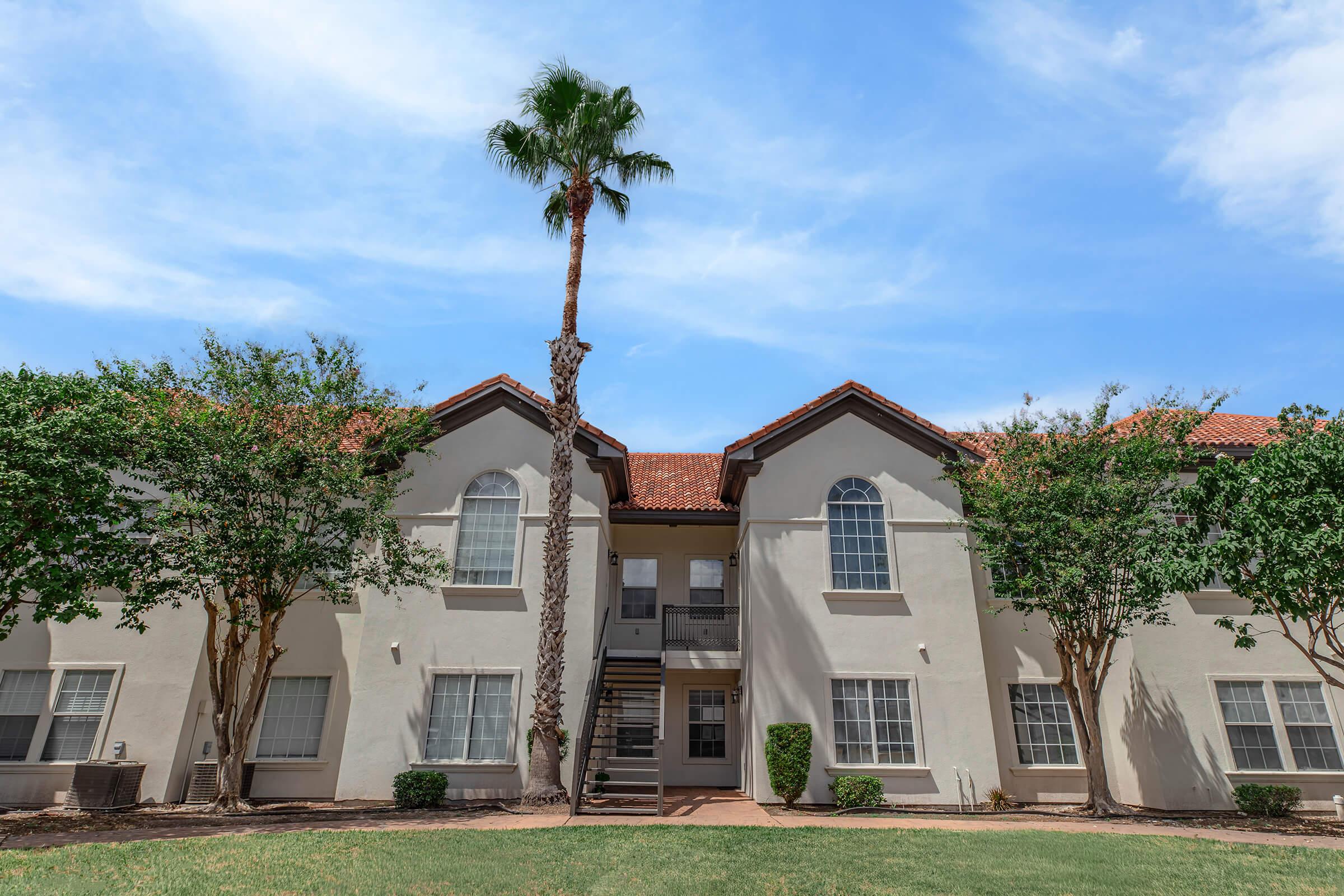 A two-story residential building with a tan facade, featuring arched windows and a red tiled roof. In front, there are well-maintained shrubs and a tall palm tree against a bright blue sky with scattered clouds. The scene conveys a warm, inviting atmosphere typical of a sunny day.