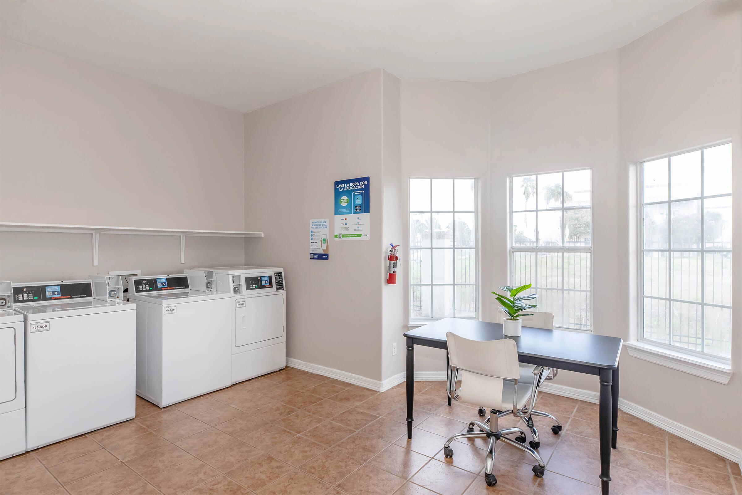 A clean laundry room featuring three washing machines and a dryer, with a small desk and chair beside large windows that let in natural light. The walls are painted in a light color, and there's a small plant on the desk, creating a pleasant and inviting atmosphere.