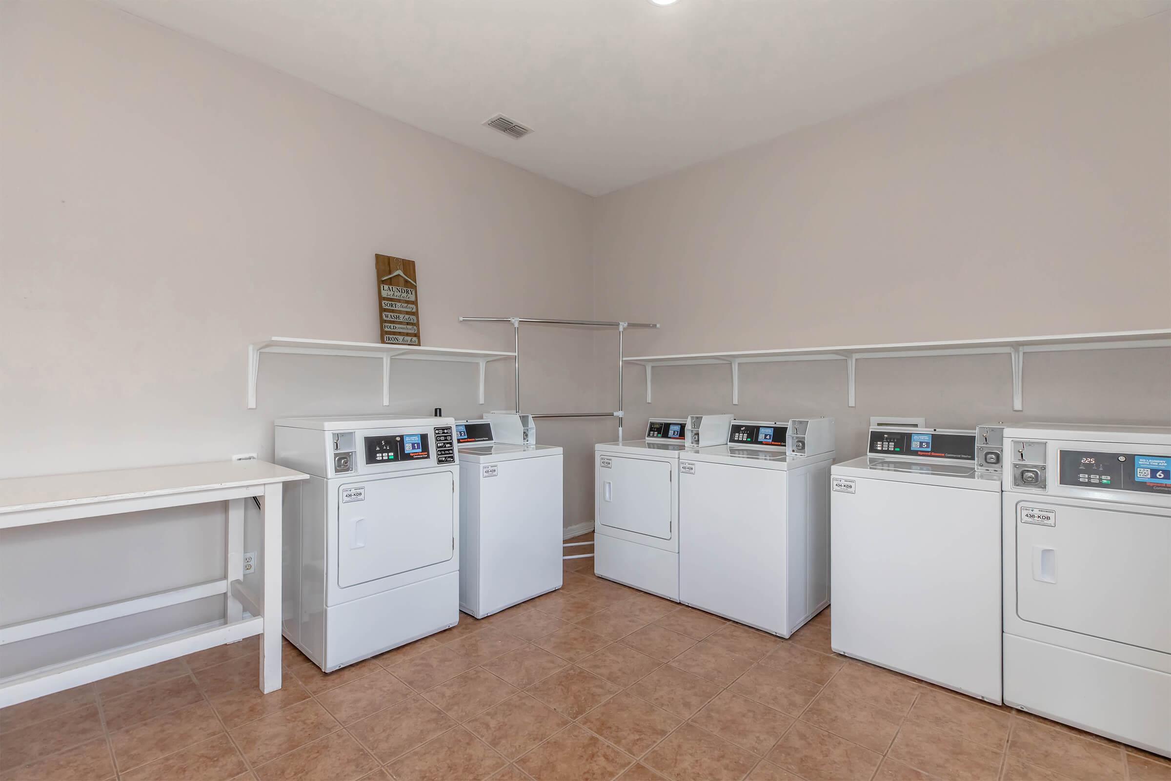A clean, modern laundry room featuring multiple white washing machines and dryers, a folding table, and a simple wall-mounted drying rack. The room has a light-colored wall and tiled flooring, creating a spacious and organized environment for laundry tasks.