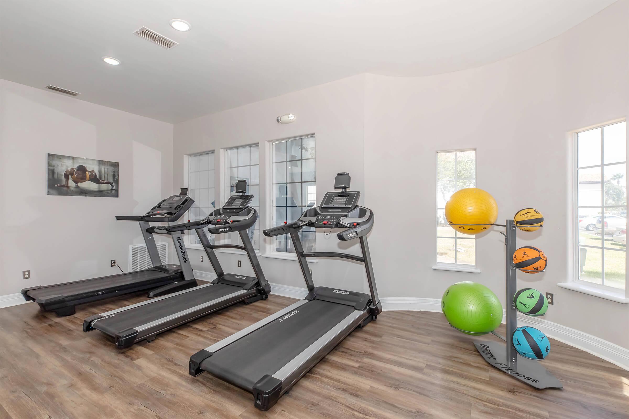 A fitness room featuring three treadmills lined up against a wall with windows. In the corner, there is a stand with colorful exercise balls. The walls are painted light, and there is a motivational image on the wall. The floor is wooden, creating a warm atmosphere for workouts.