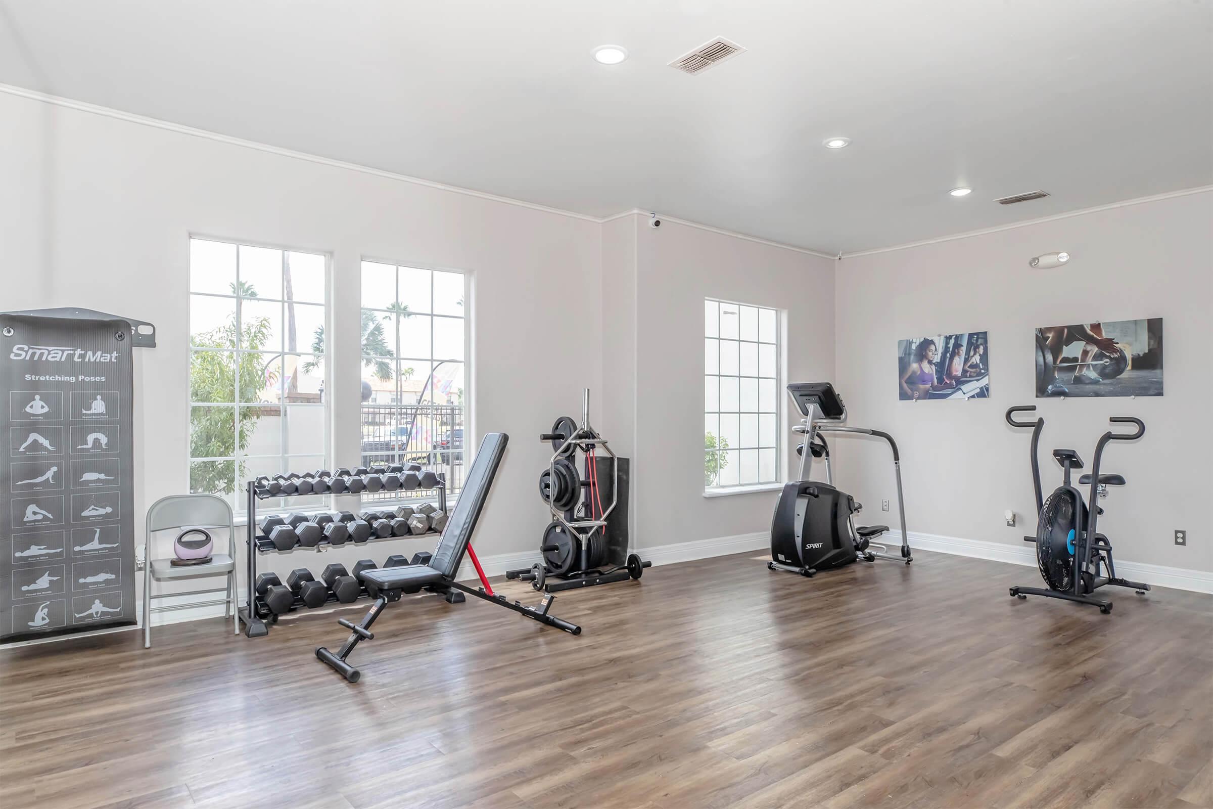 A well-lit fitness room featuring a variety of exercise equipment. On the left, a rack of dumbbells is visible next to a multi-use weight bench. In the center, a treadmill and an elliptical machine are positioned near large windows, allowing natural light to fill the space. Two framed workout images adorn the walls.