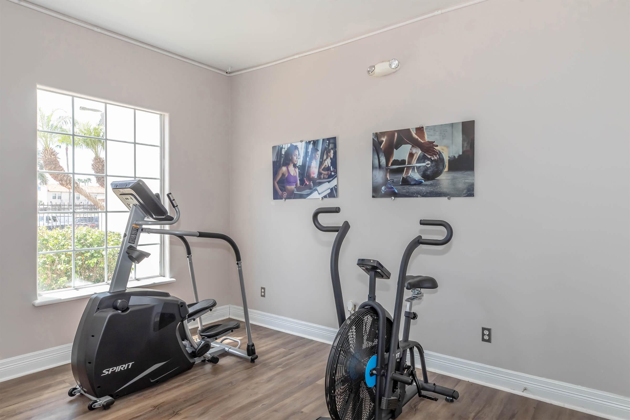 A fitness room with exercise equipment including a stationary bike and an air bike. The walls are adorned with two motivational fitness-themed pictures. A large window allows natural light to brighten the space, creating an inviting atmosphere for workouts.