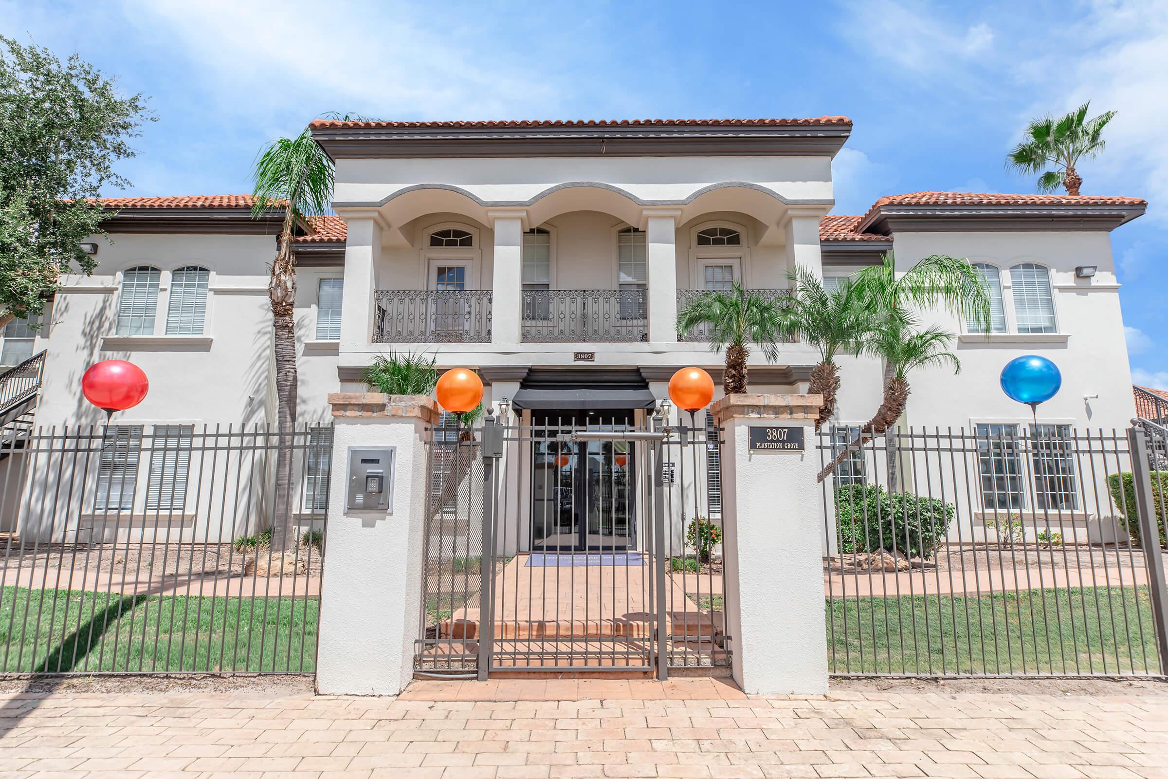A charming two-story building with a modern facade, featuring a gated entrance. The yard is accented with greenery and two large colorful balloons—one orange and one blue—flanking the entry. The sky is clear with a few clouds, enhancing the inviting atmosphere of the property.