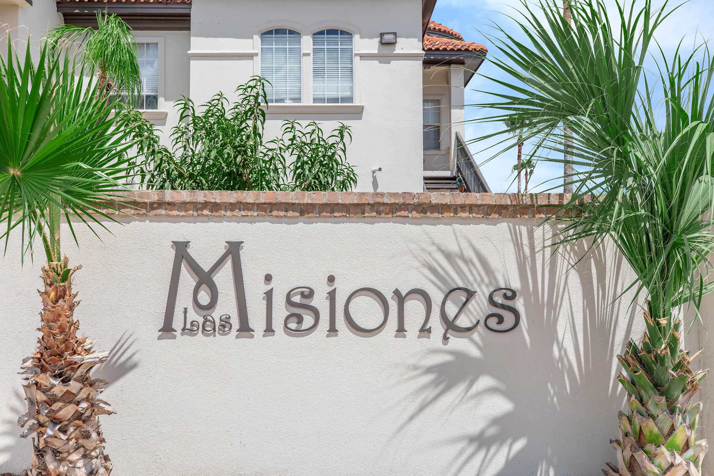 A decorative wall with the text "Las Misiones" prominently displayed. Surrounding the wall are green palm trees, and a light-colored building is partially visible in the background with a blue sky above.